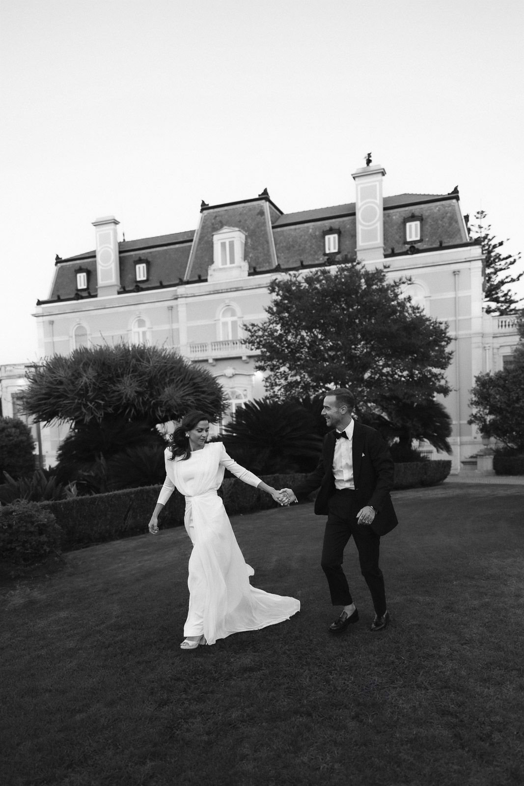 Black and white photo of a bride and groom holding hands and dancing on a lawn in front of an elegant mansion.