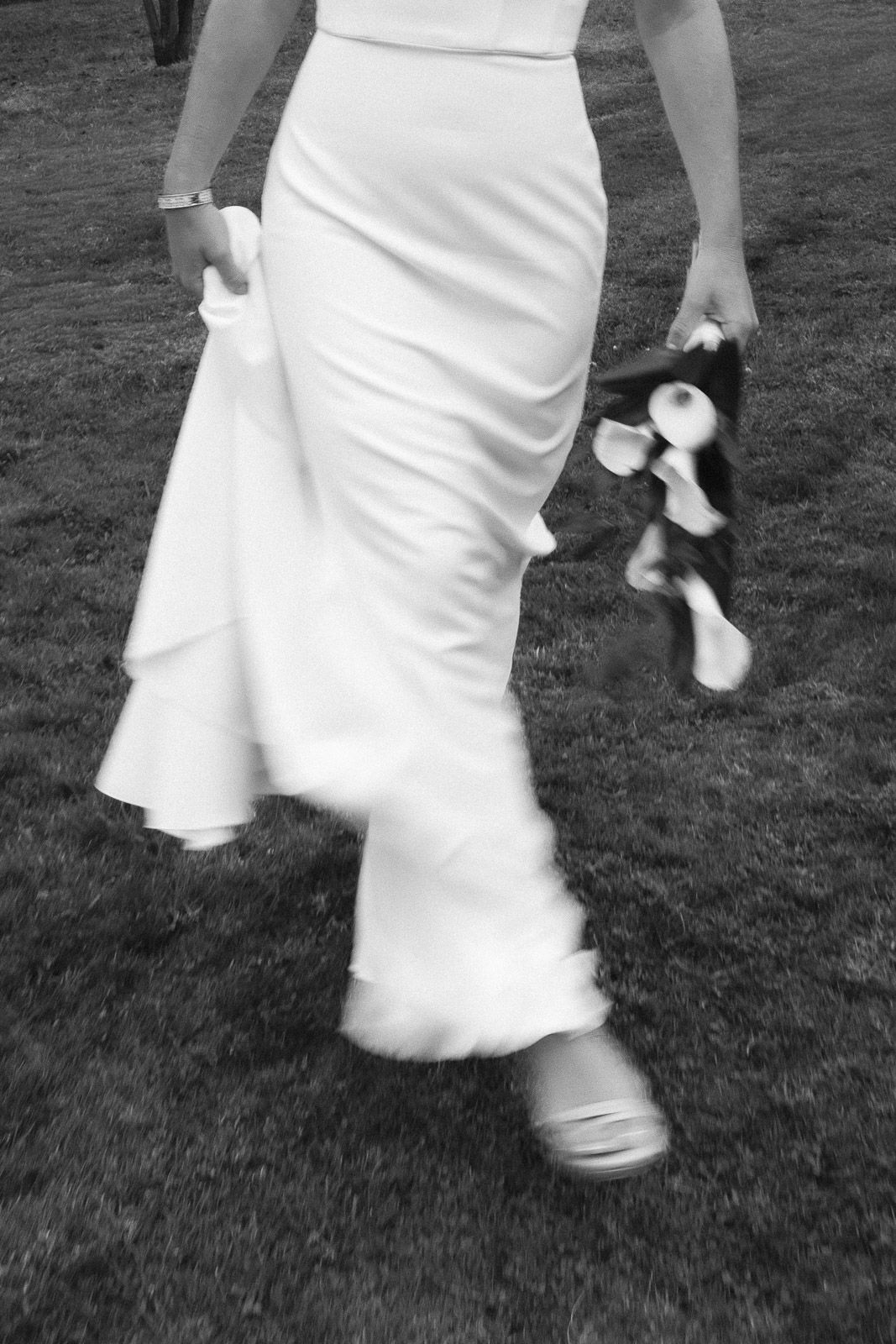 Black and white photo of a bride in a white gown holding a bouquet, walking across a grass lawn.