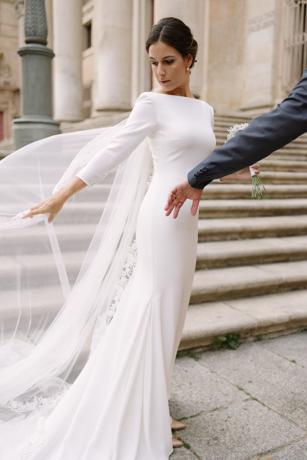 Elegant bride in a fitted white long-sleeve wedding gown with flowing veil on stone steps.
