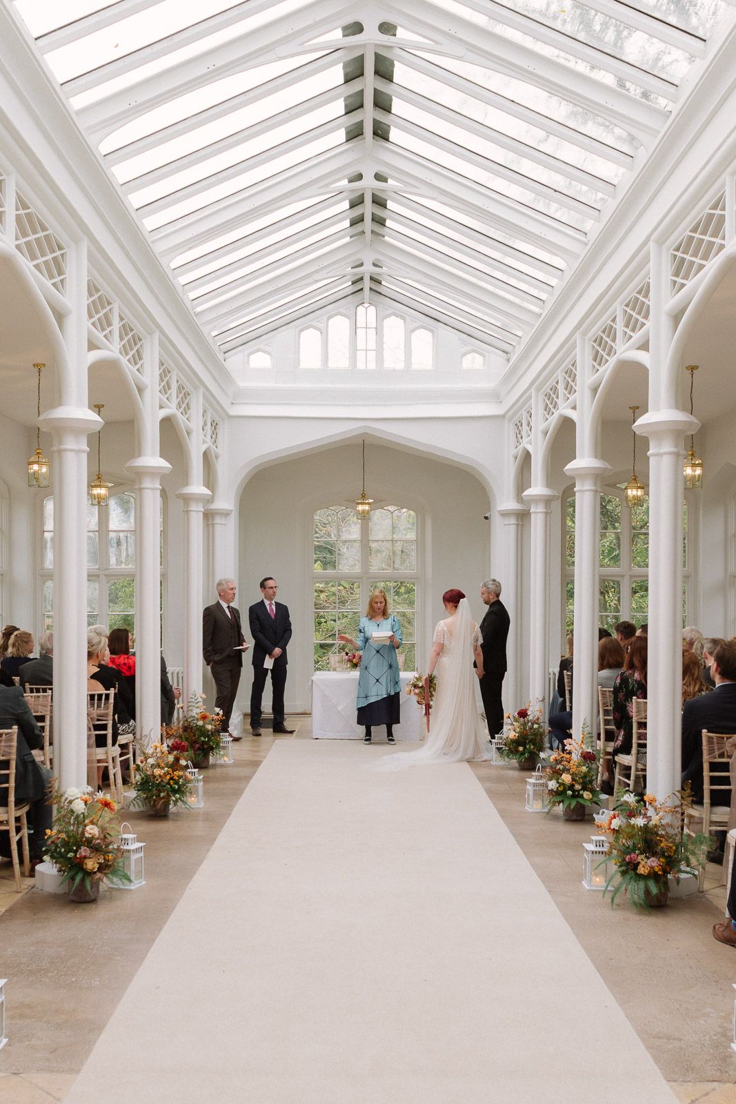 Elegant wedding ceremony in a bright glass-roofed conservatory with white columns, floral arrangements, and guests seated along the aisle.