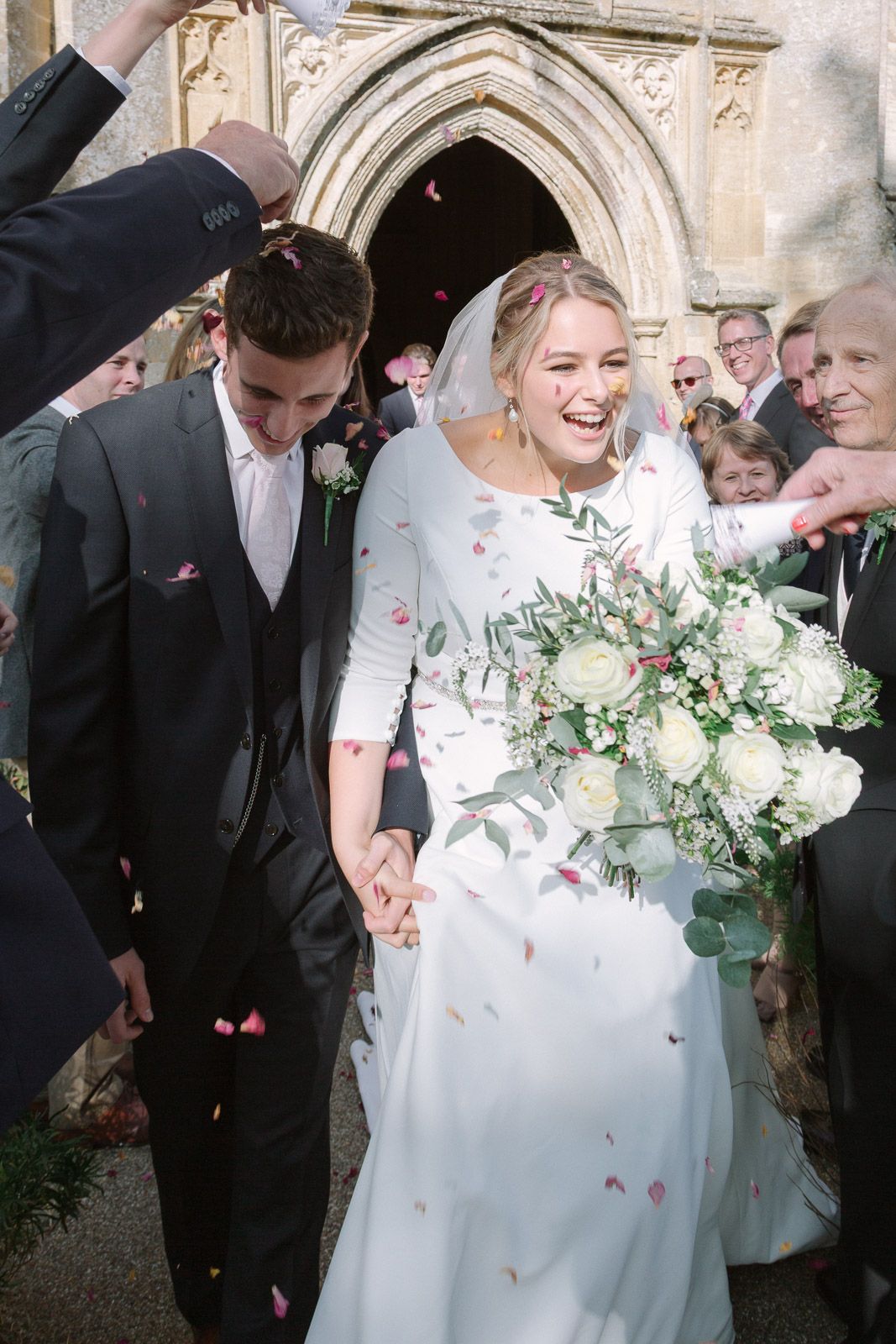 Bride and groom exit church amid confetti shower, bride holding white rose bouquet, guests celebrating wedding ceremony.