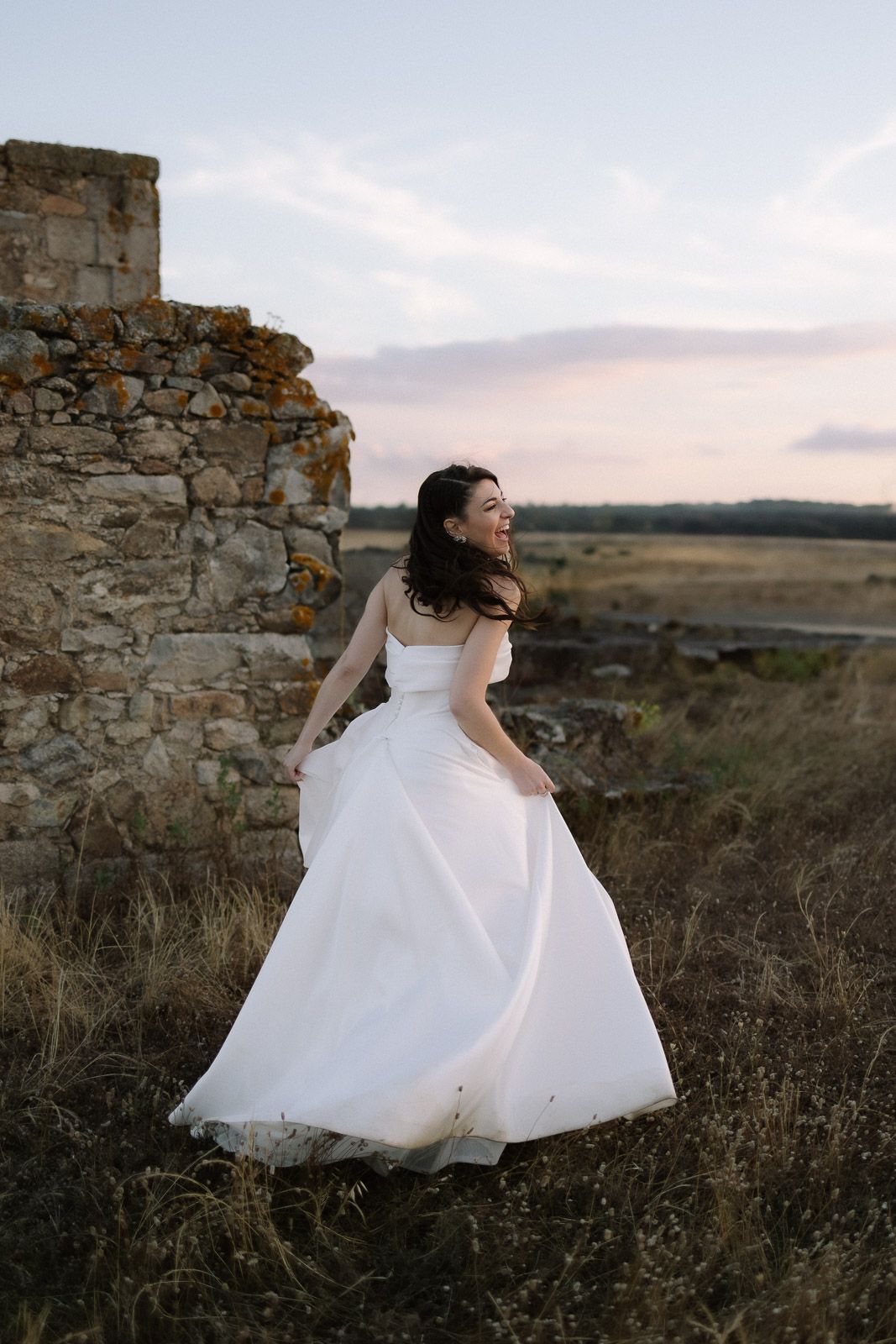 Smiling bride in white gown twirling near stone ruins at sunset in an open field.