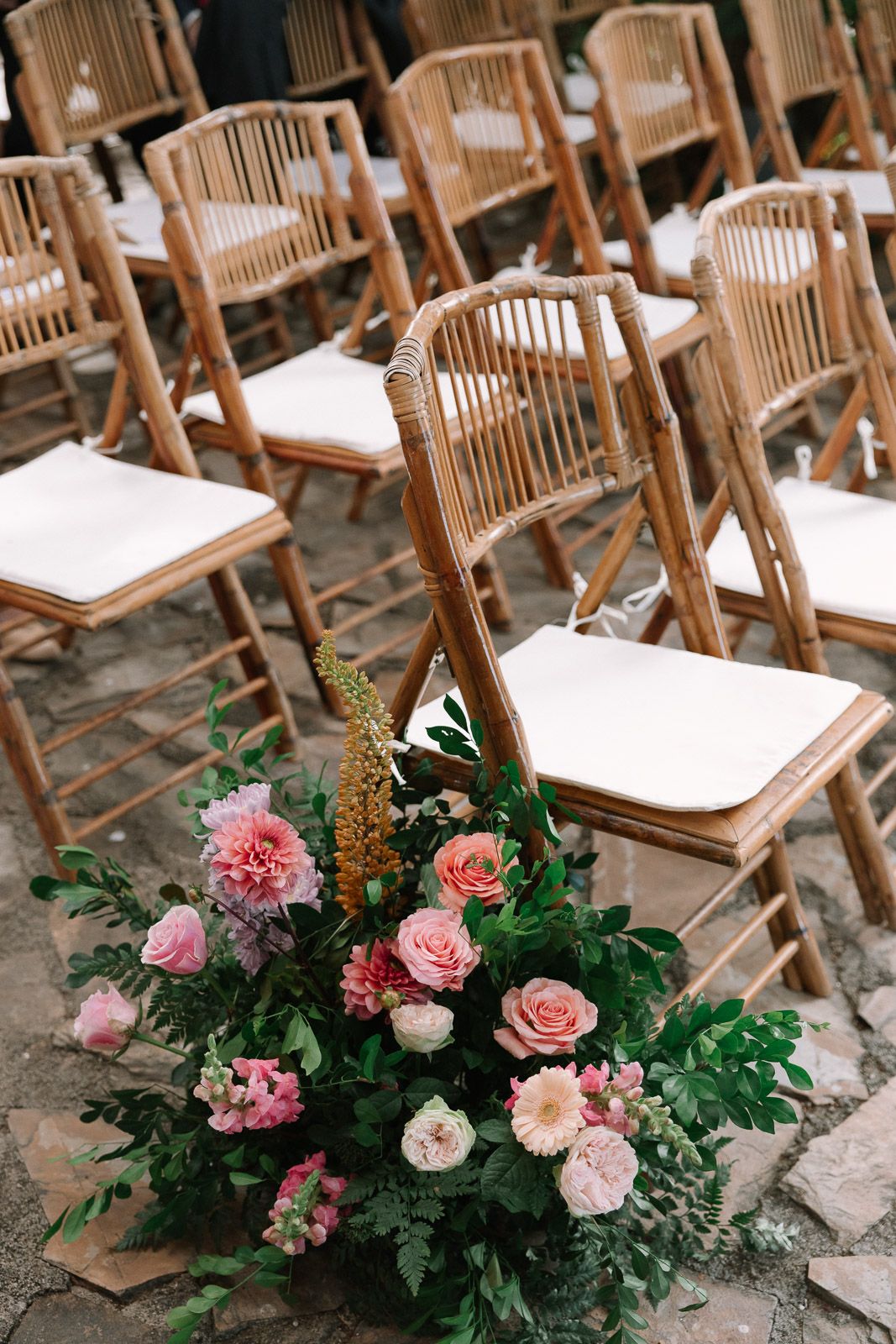 Bamboo folding chairs with white cushions arranged for an outdoor wedding ceremony with pink floral arrangement on stone floor.