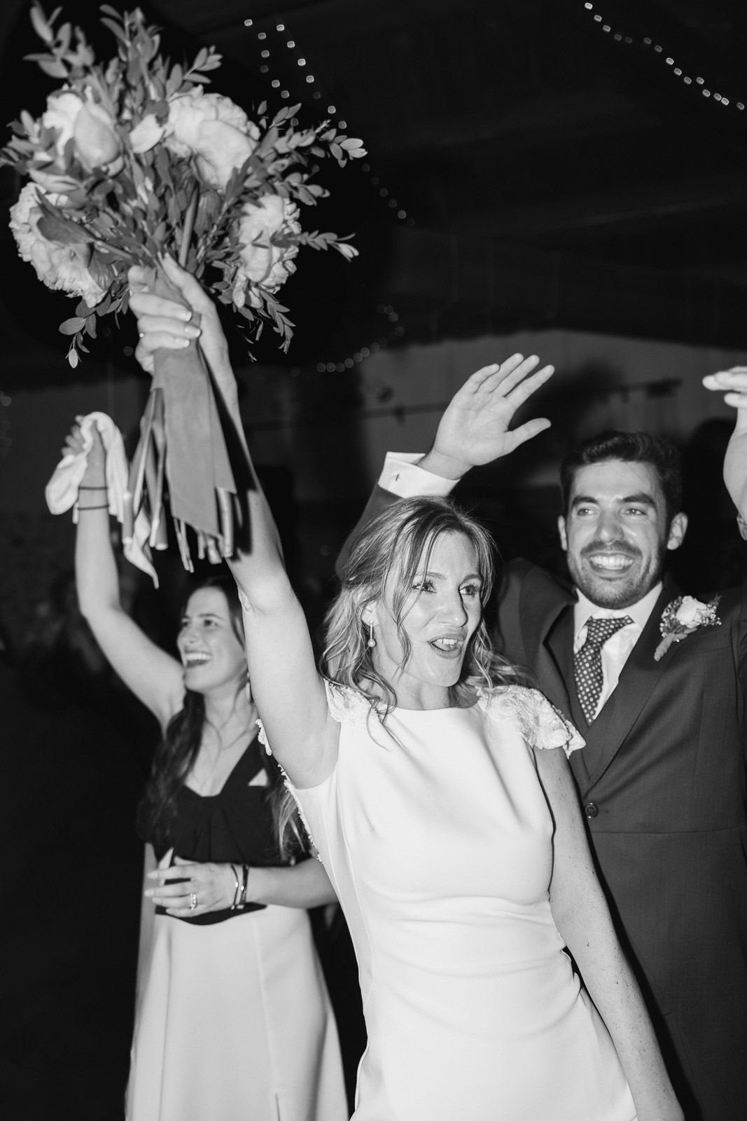 Black and white photo of a joyful bride in white dress raising bouquet with groom and guests celebrating at wedding reception.