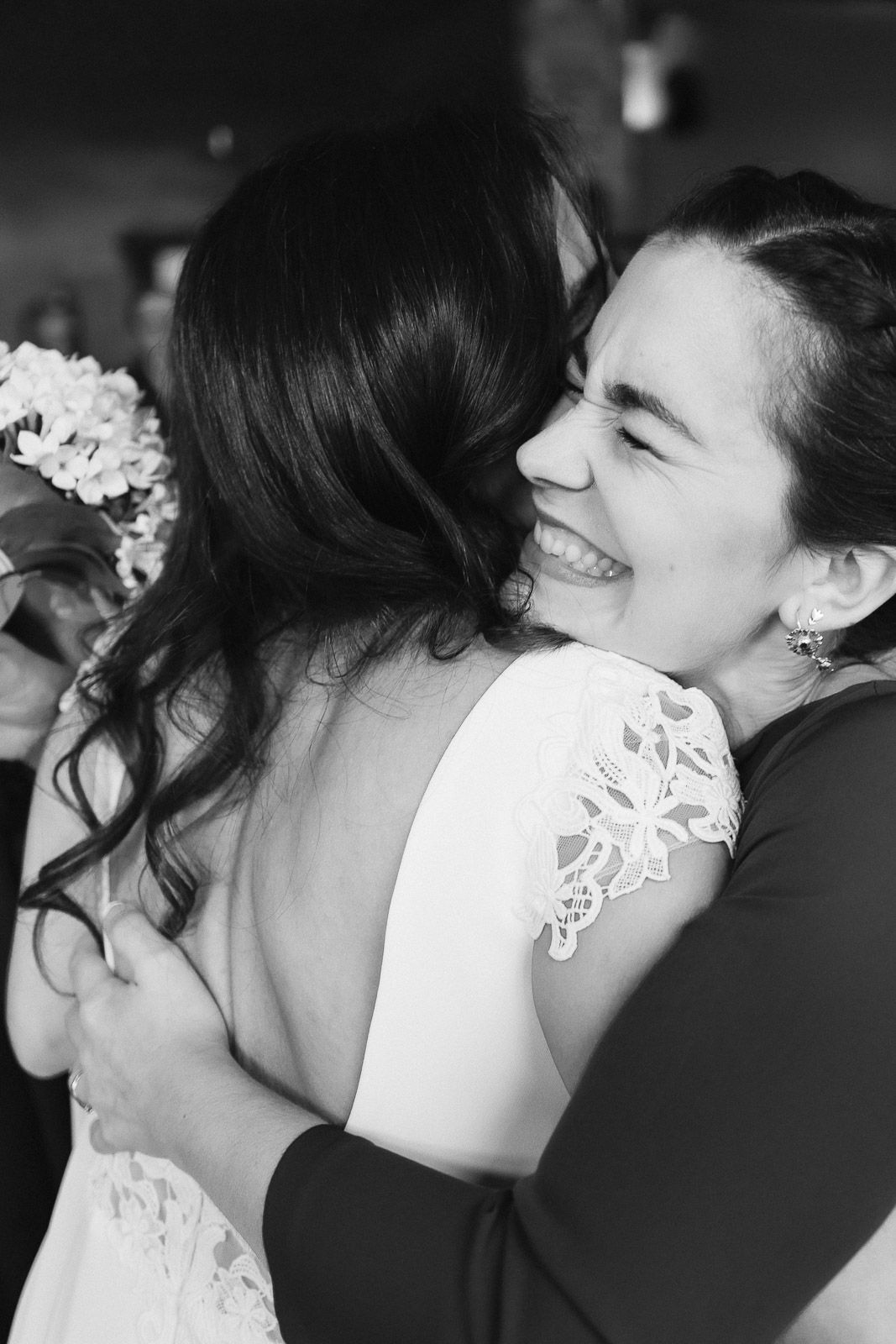 Black and white photo of two women sharing a joyful hug at a wedding, bride in lace dress smiling warmly.