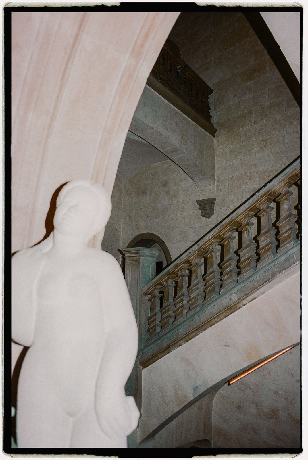 White marble statue beside an ornate staircase with balustrade in a grand classical building interior.