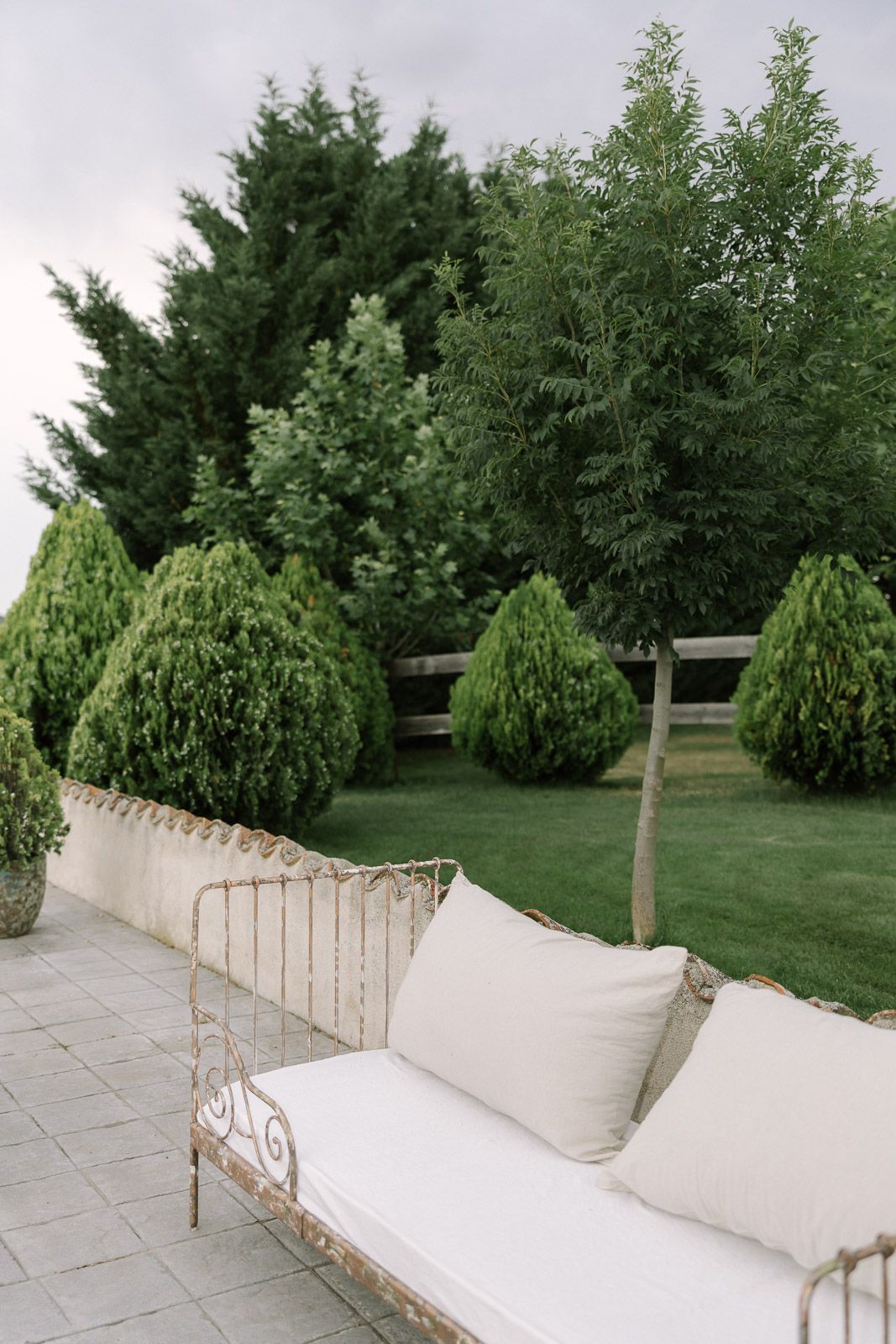 Elegant white iron garden sofa with cushions on a stone patio surrounded by lush green topiary trees.