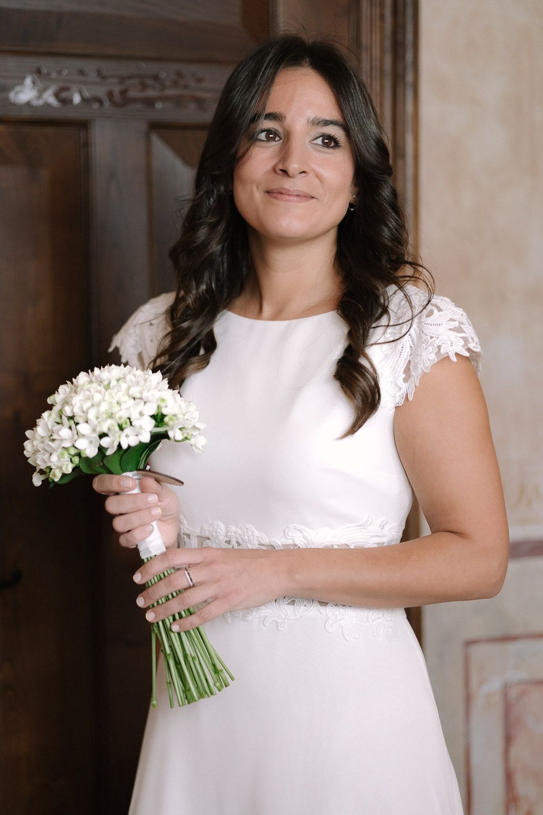 Bride in white lace cap-sleeve gown holding white floral bouquet, smiling indoors near wooden door.