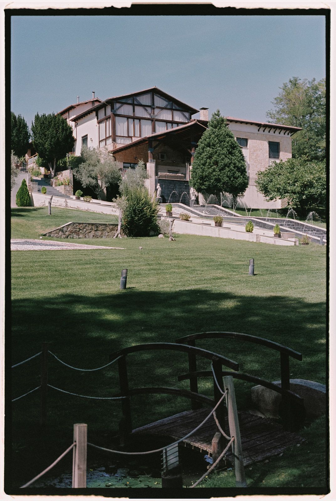 Rustic estate with wooden bridge over pond, manicured lawns, and traditional architecture surrounded by lush greenery.