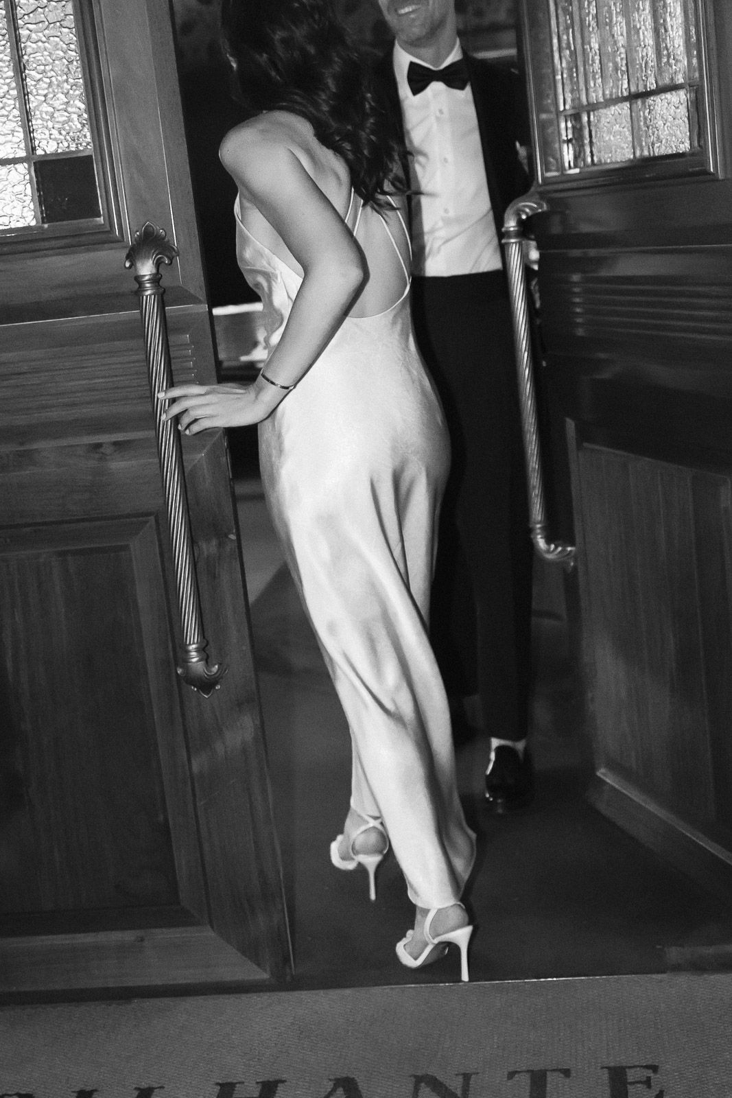 Elegant couple in formal attire entering a venue, woman in backless white gown and heels, man in tuxedo.