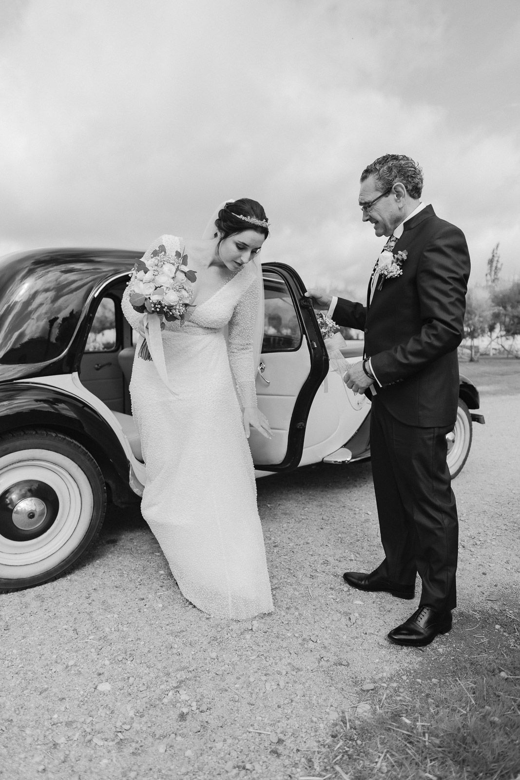 Black and white photo of a groom helping his bride exit a vintage car on their wedding day.