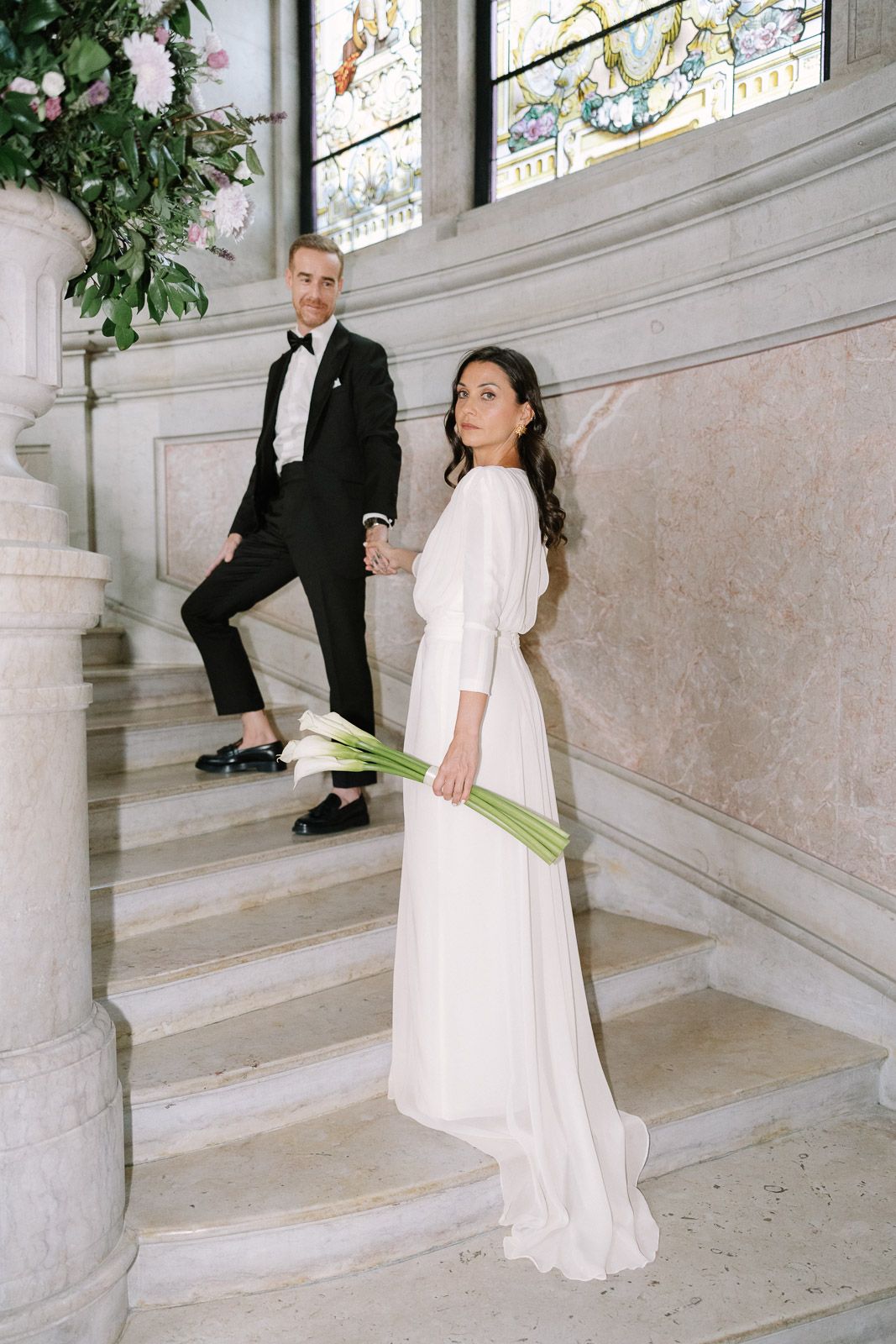 Bride in white gown holds calla lilies on marble stairs beside groom in black tuxedo in elegant venue.