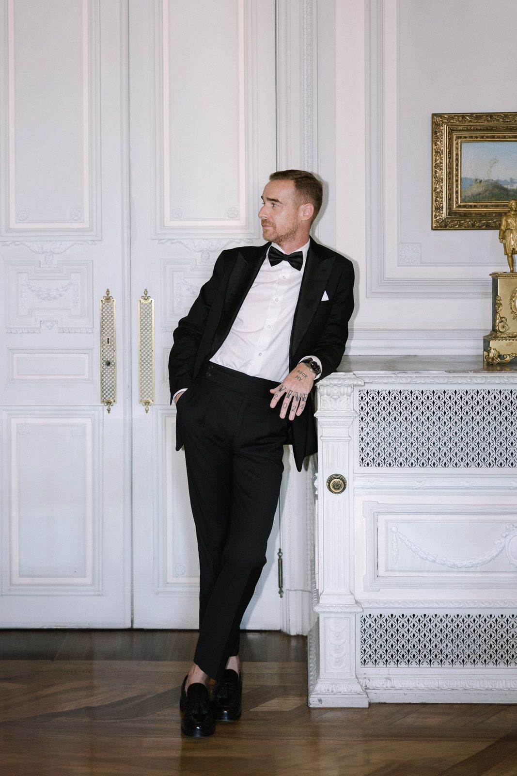 Elegant groom in black tuxedo and bow tie leaning against white ornate wall in luxury venue.