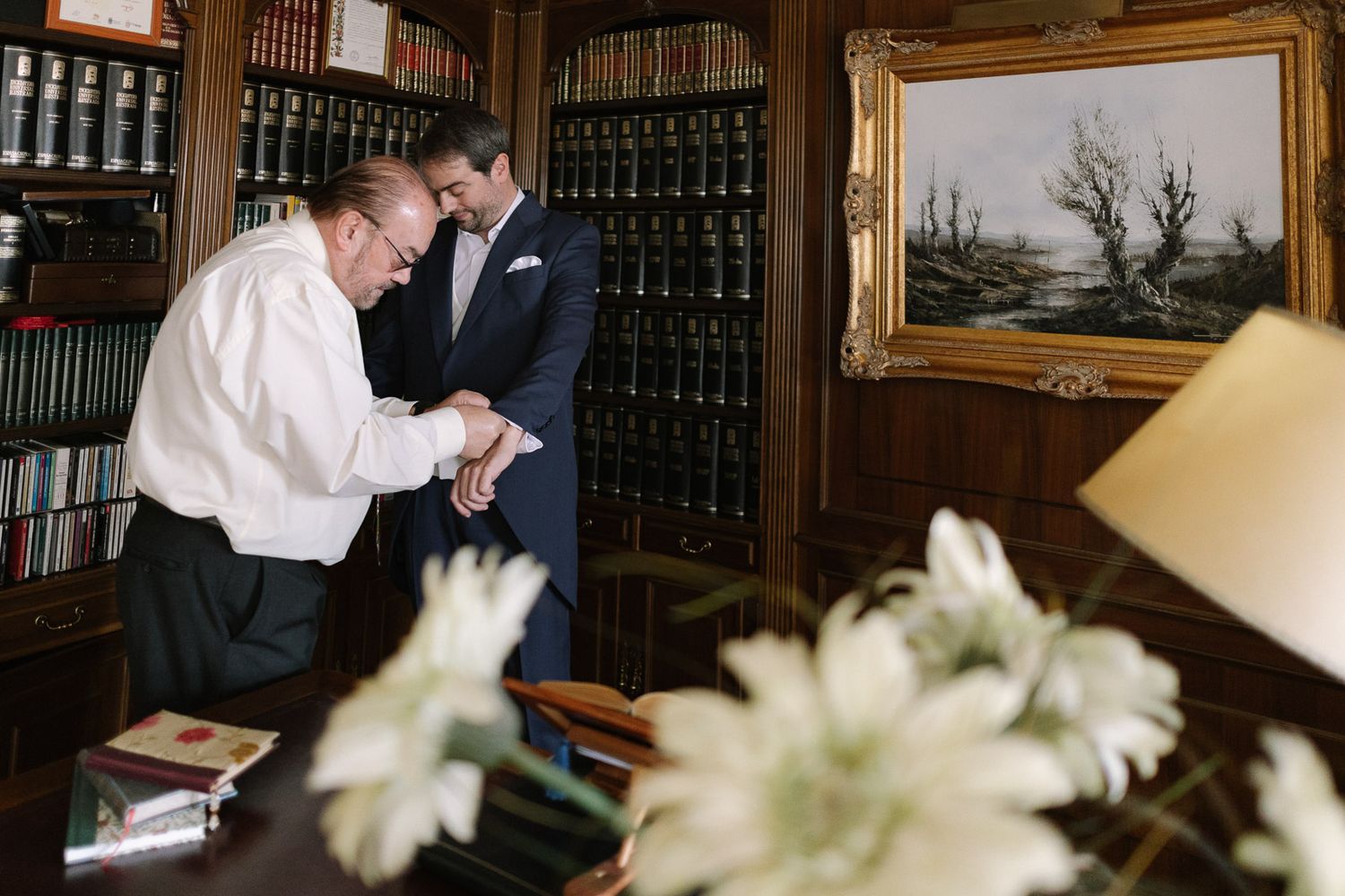 Two men in formal attire examine documents in an elegant wood-paneled library with bookshelves and classic artwork.