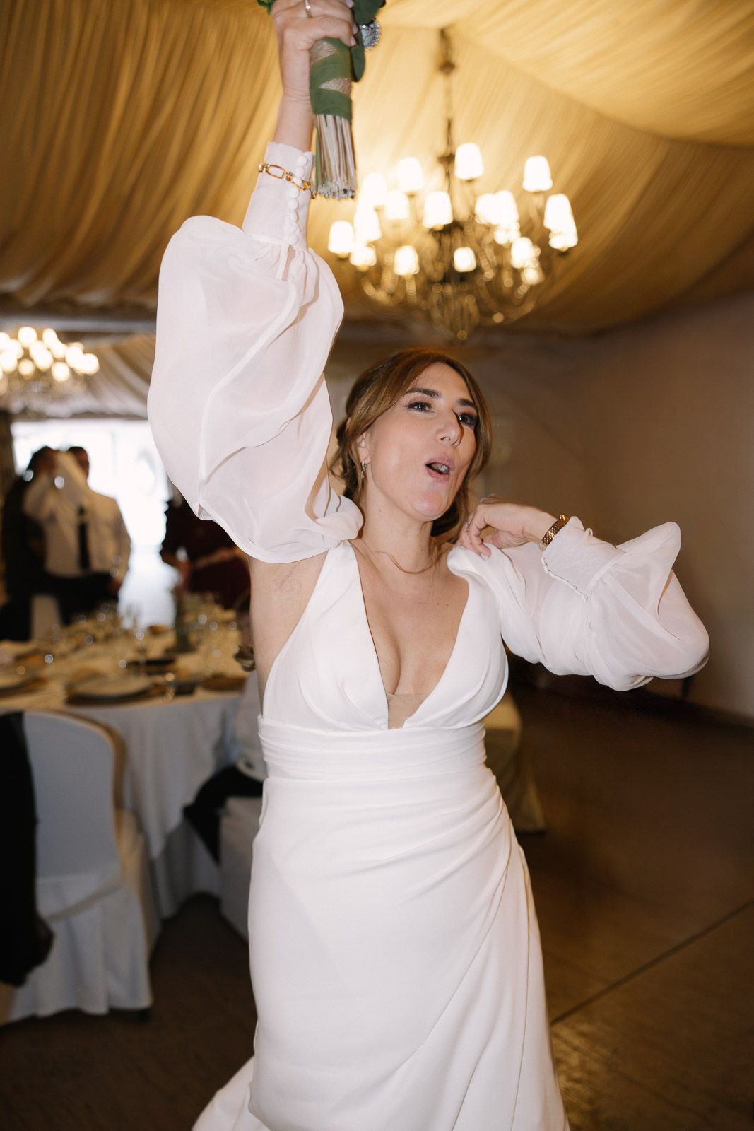 Bride in white gown with puffed sleeves raises bouquet at elegant wedding reception venue.