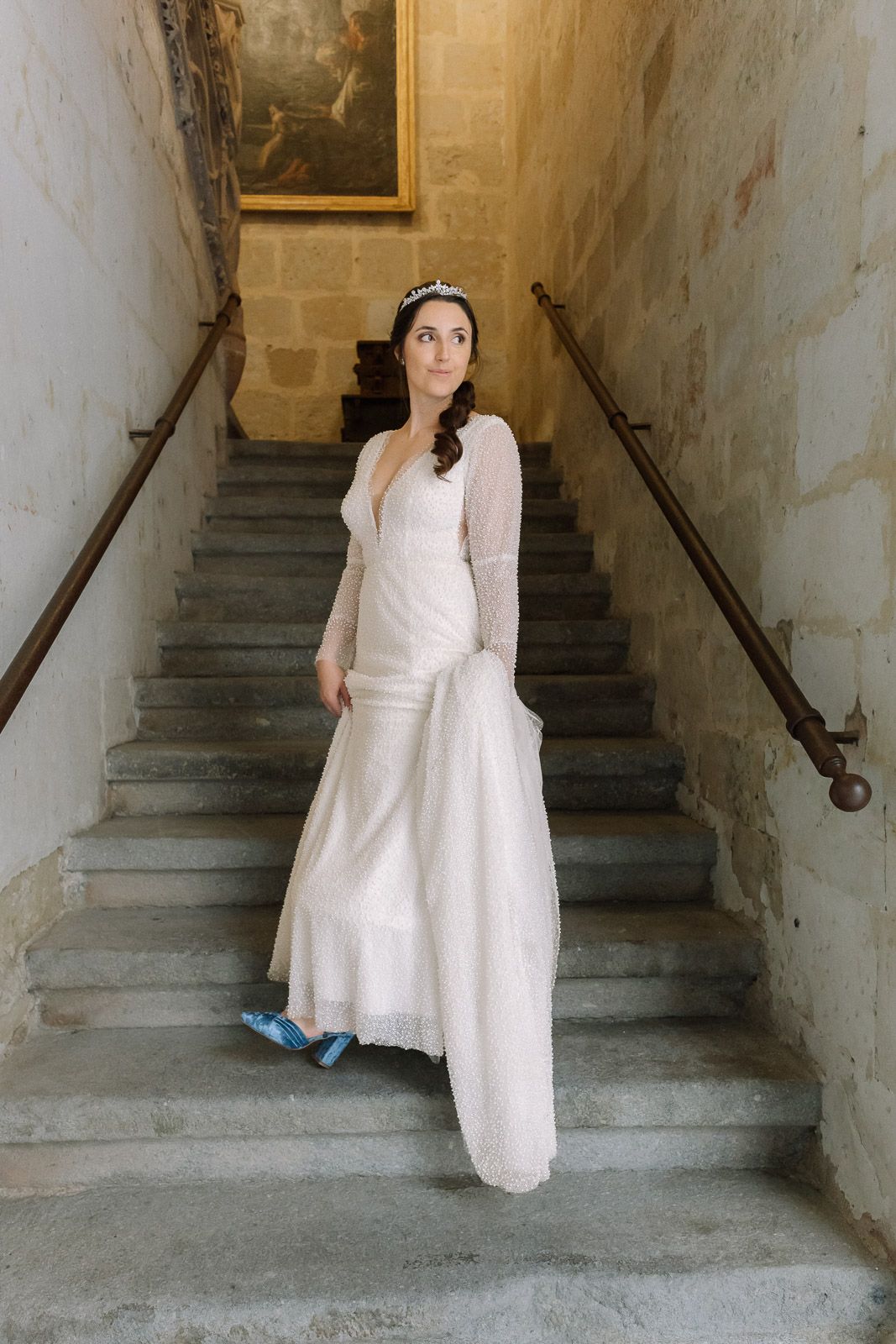 Bride in white wedding gown and blue shoes stands on stone staircase wearing tiara, looking up gracefully.