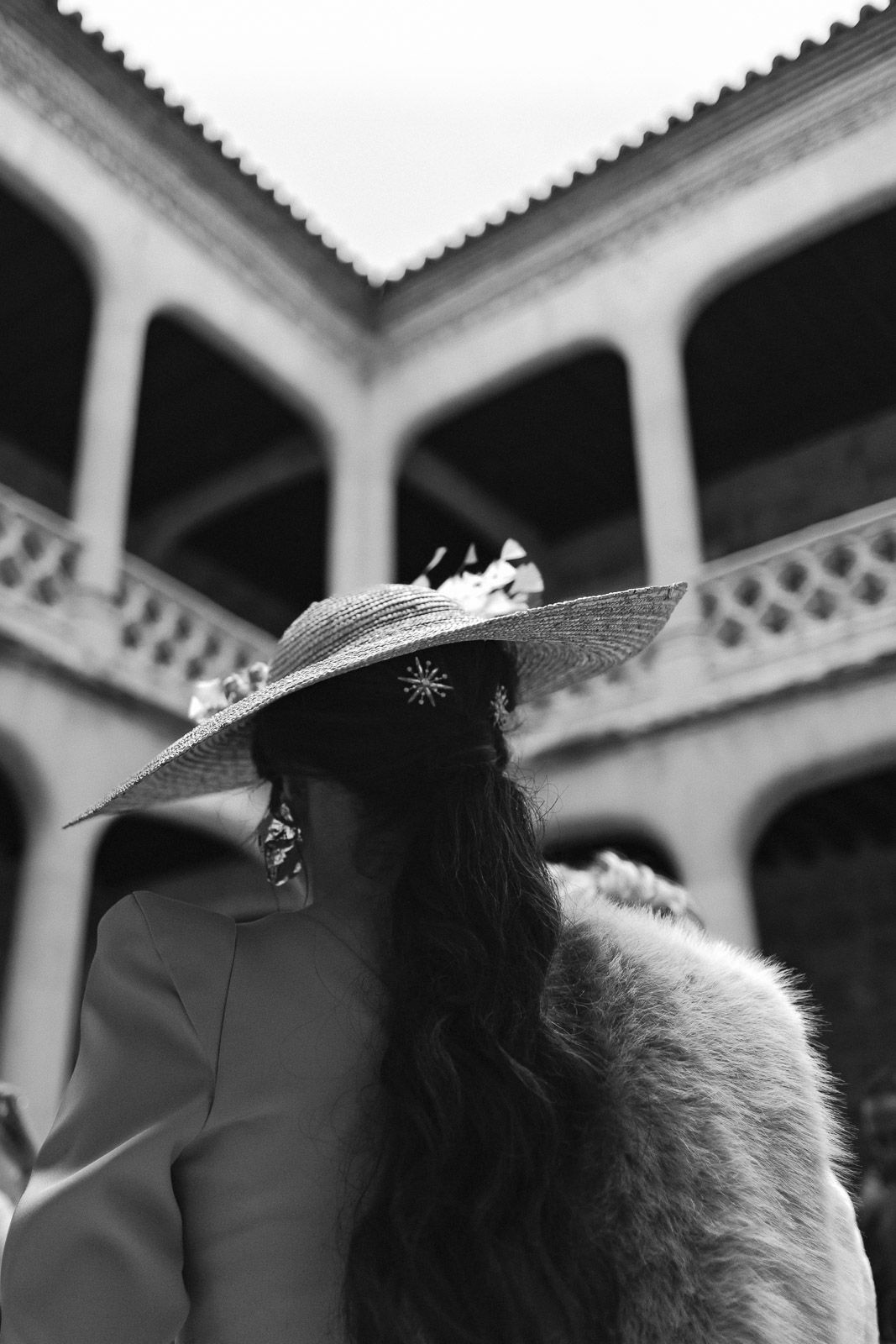 Black and white photo of a woman in a wide-brim hat with star pin and fur stole before an ornate building.