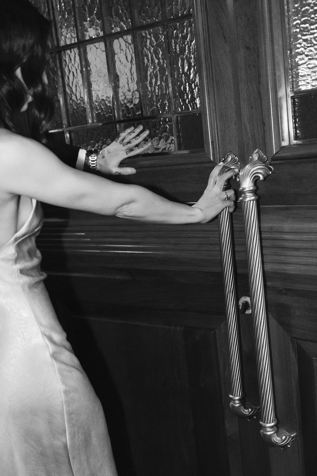 Woman in silk slip dress pushing open ornate brass door handles in black and white photo.