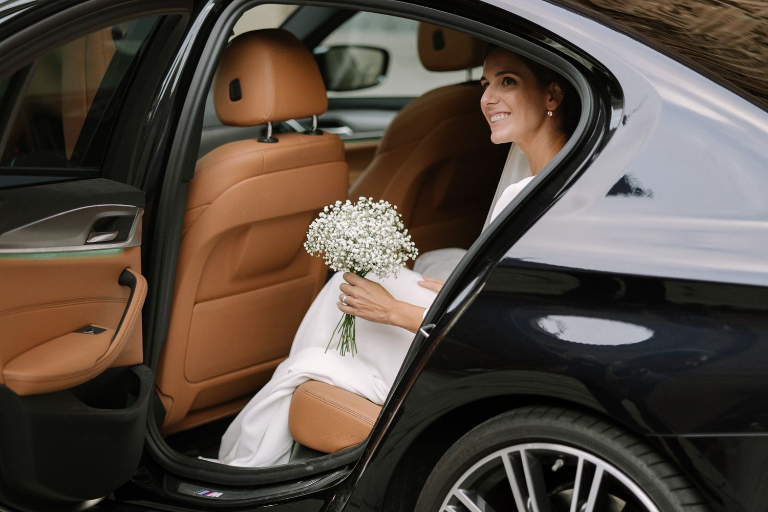 Smiling bride in white dress holding white flower bouquet sitting in luxury car with tan leather interior.