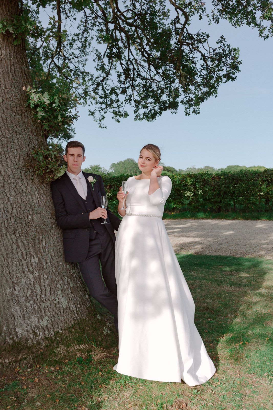 Bride and groom holding champagne glasses leaning against a large tree outdoors on their wedding day.