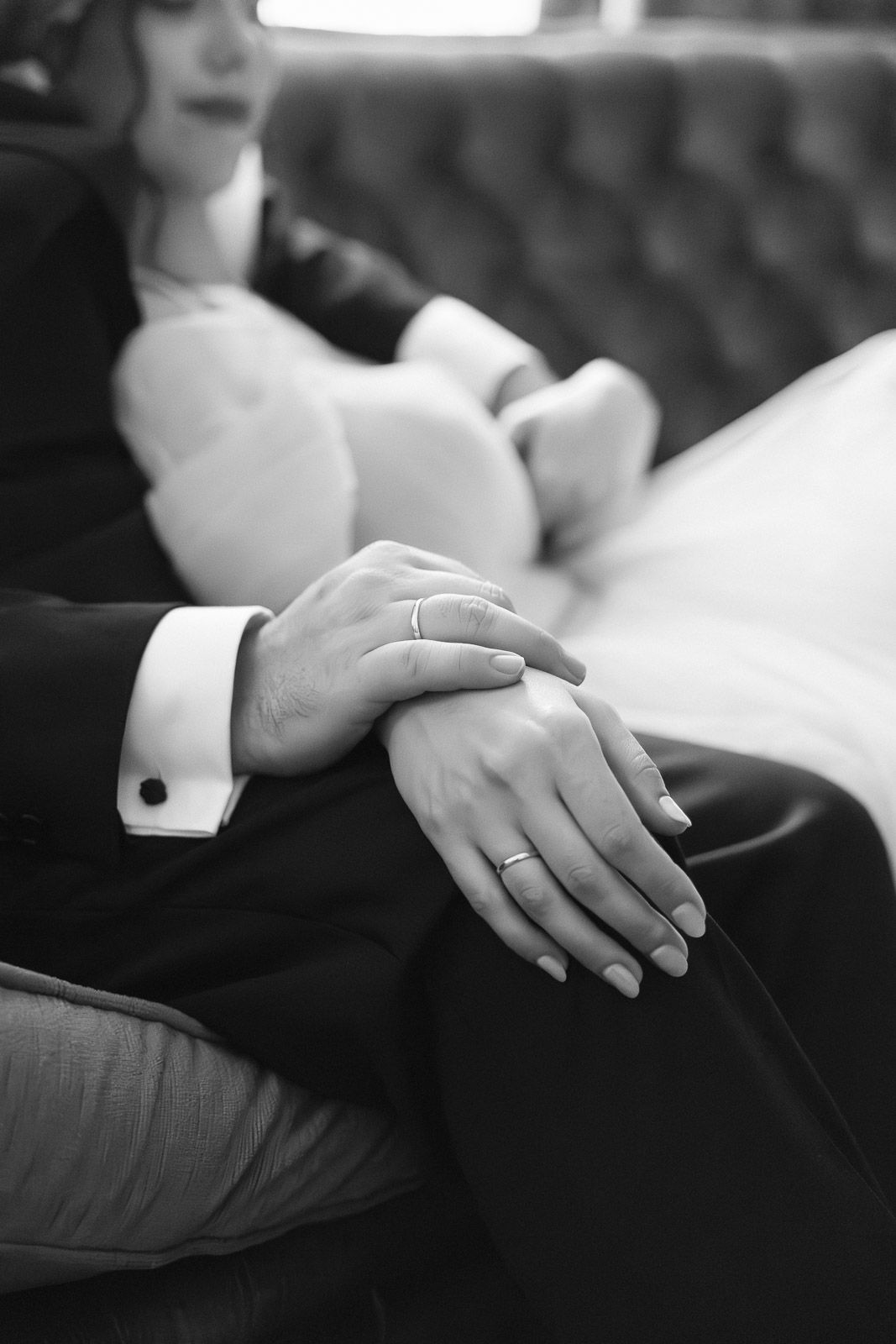 Black and white photo of newlyweds hands with wedding rings resting together on grooms suit.