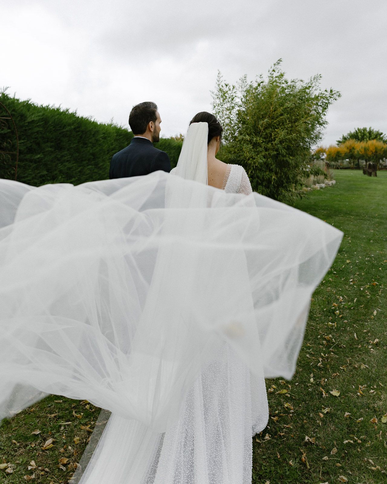 A bride and groom walk through lush gardens, her flowing white veil billowing dramatically in the wind.