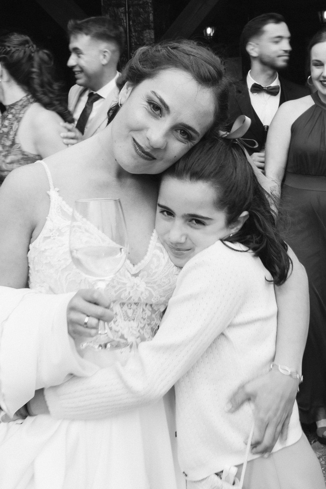 Black and white photo of a bride in lace dress hugging a young girl at a wedding reception.