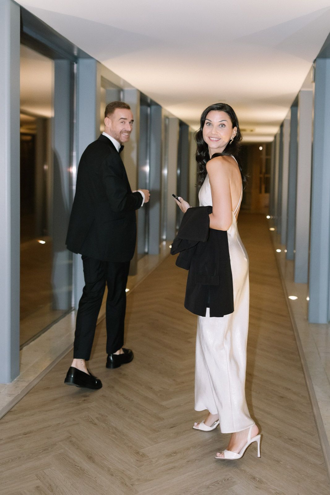 Elegant couple in formal attire walking down a modern hotel corridor, woman in white dress, man in black suit.
