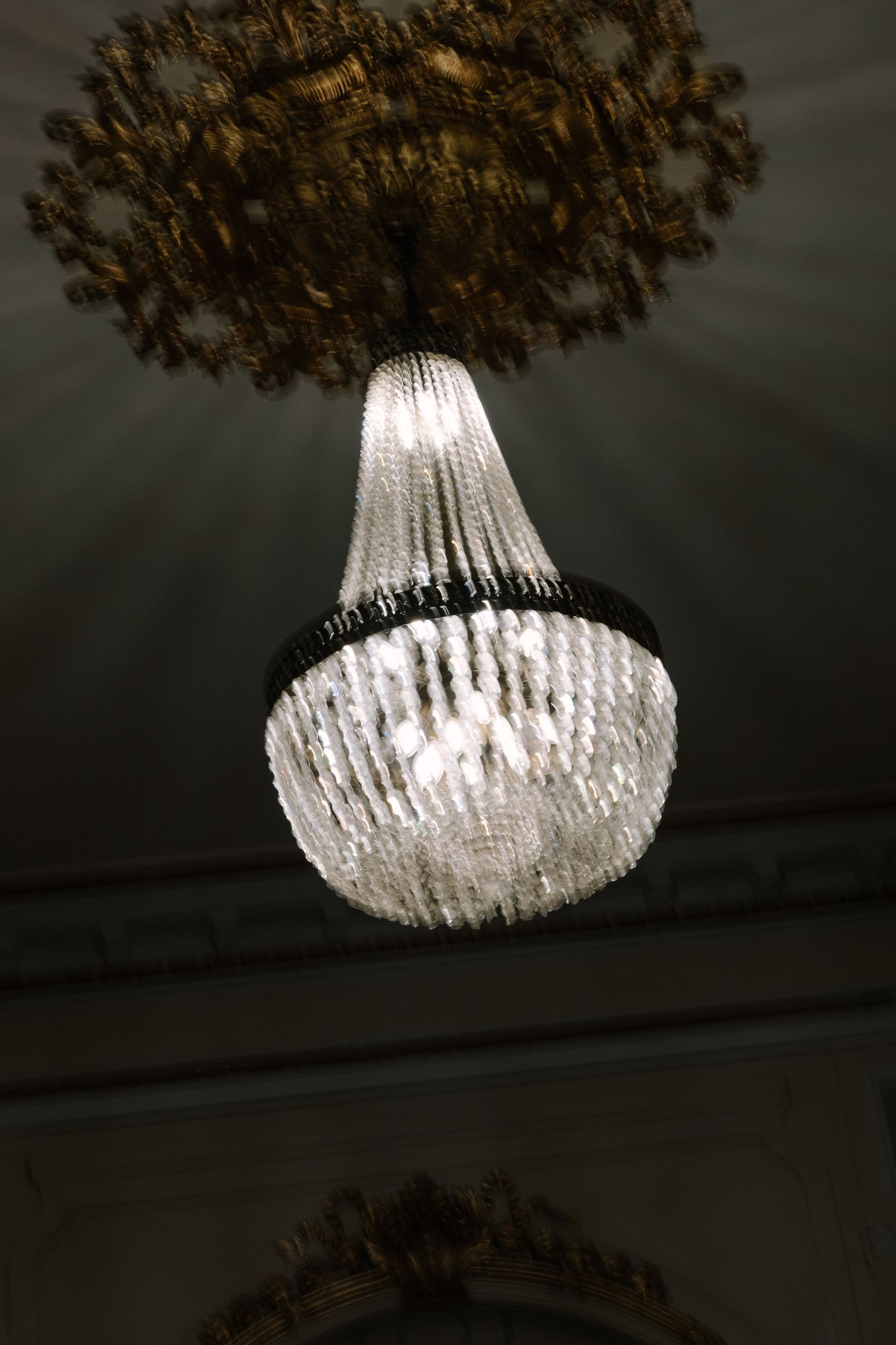 An elegant crystal chandelier with a white bell-shaped shade and ornate bronze ceiling mount glows against a dark background.