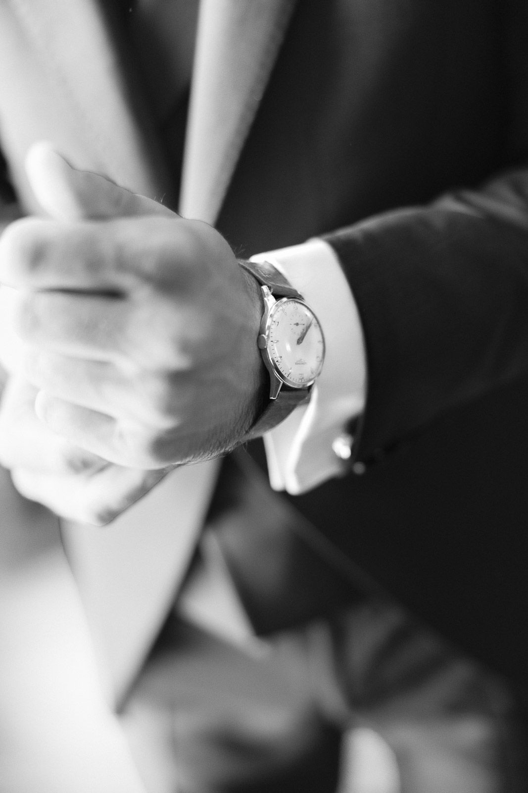 Black and white close-up of a man in a suit checking a classic analog wristwatch on his left hand.