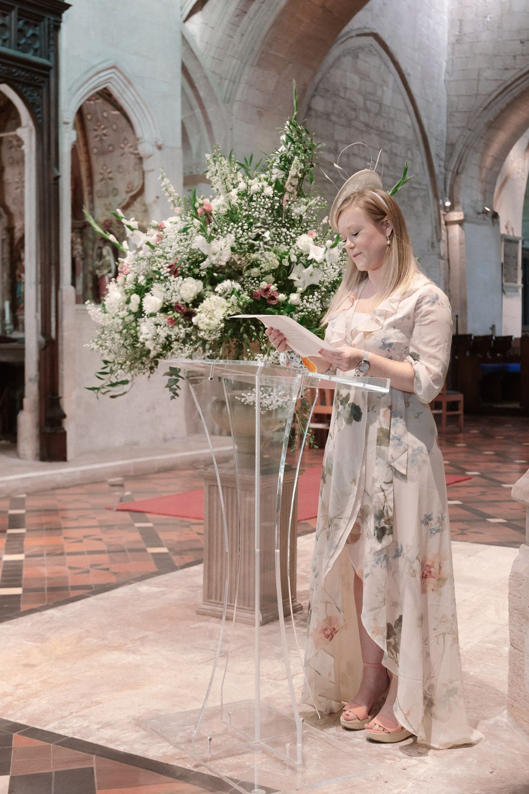 Woman in floral dress reading at clear acrylic podium adorned with white flower arrangement inside historic church.