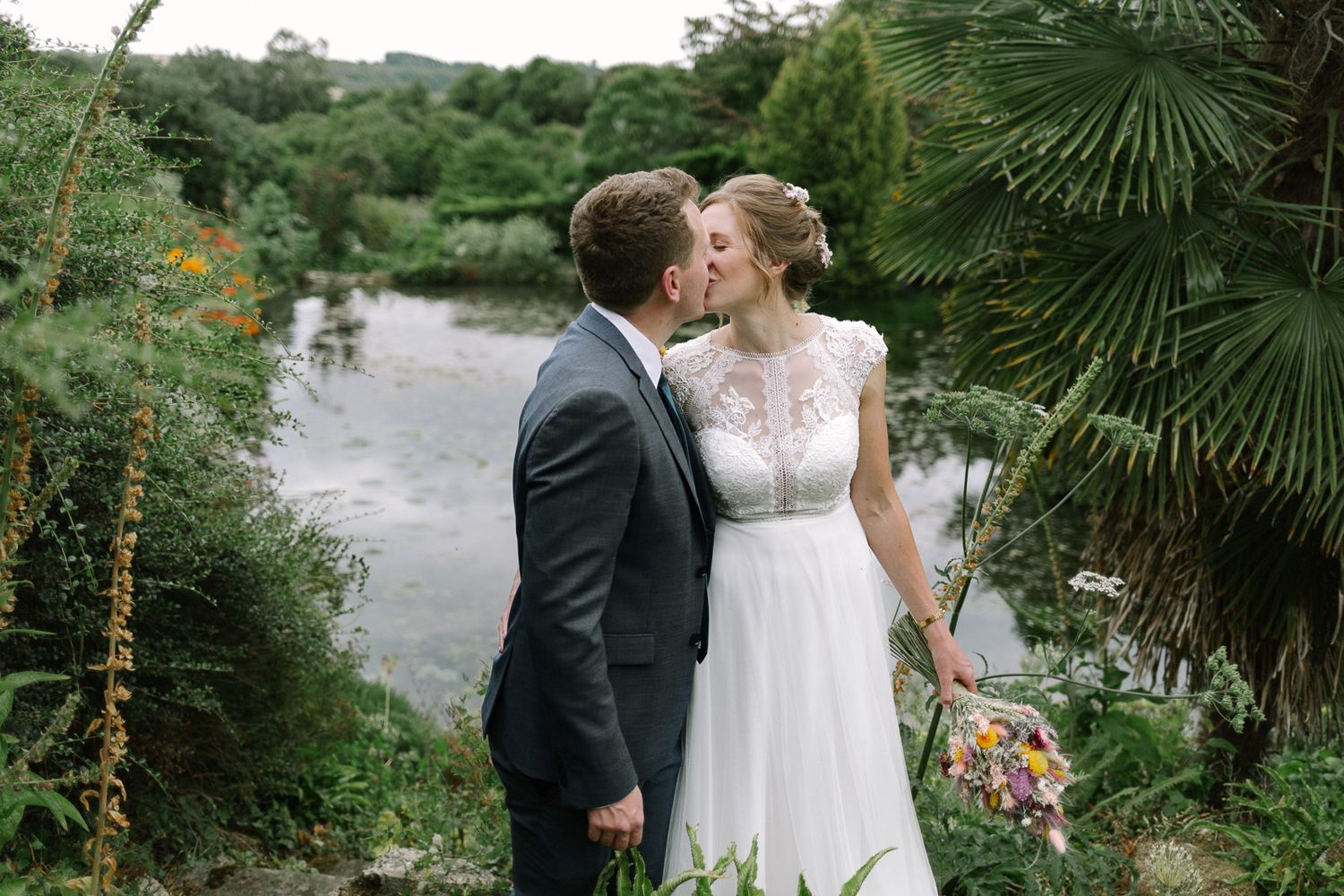 Bride and groom share a romantic kiss surrounded by lush greenery and a serene river backdrop on their wedding day.