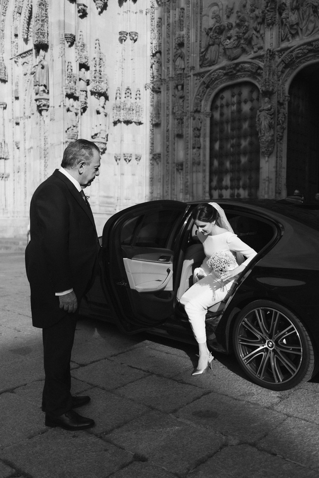Black and white photo of a bride stepping out of a luxury car assisted by a man in a tuxedo outside a grand cathedral.