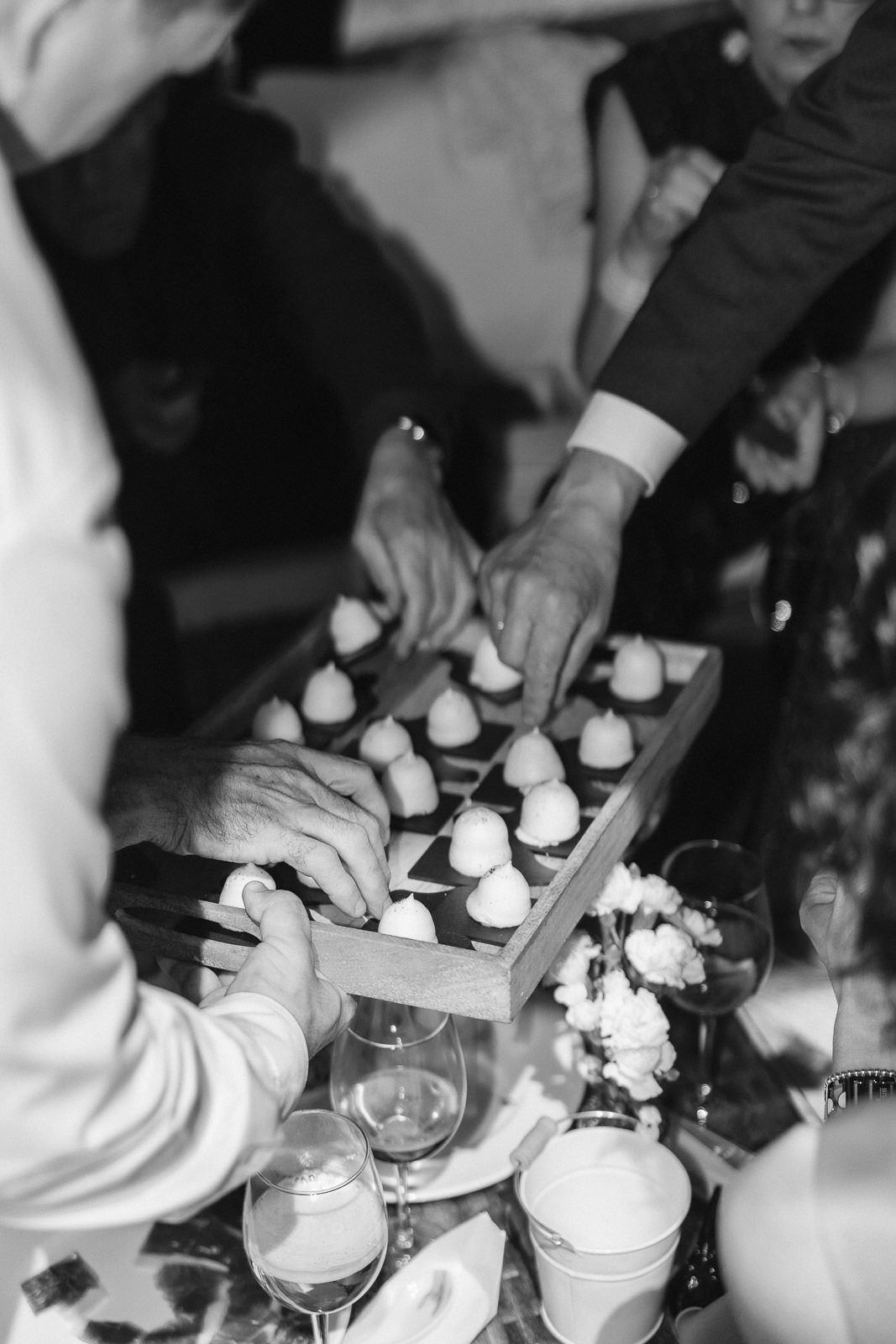 Black and white photo of guests reaching for elegant bite-sized appetizers on a wooden tray at a catered event.