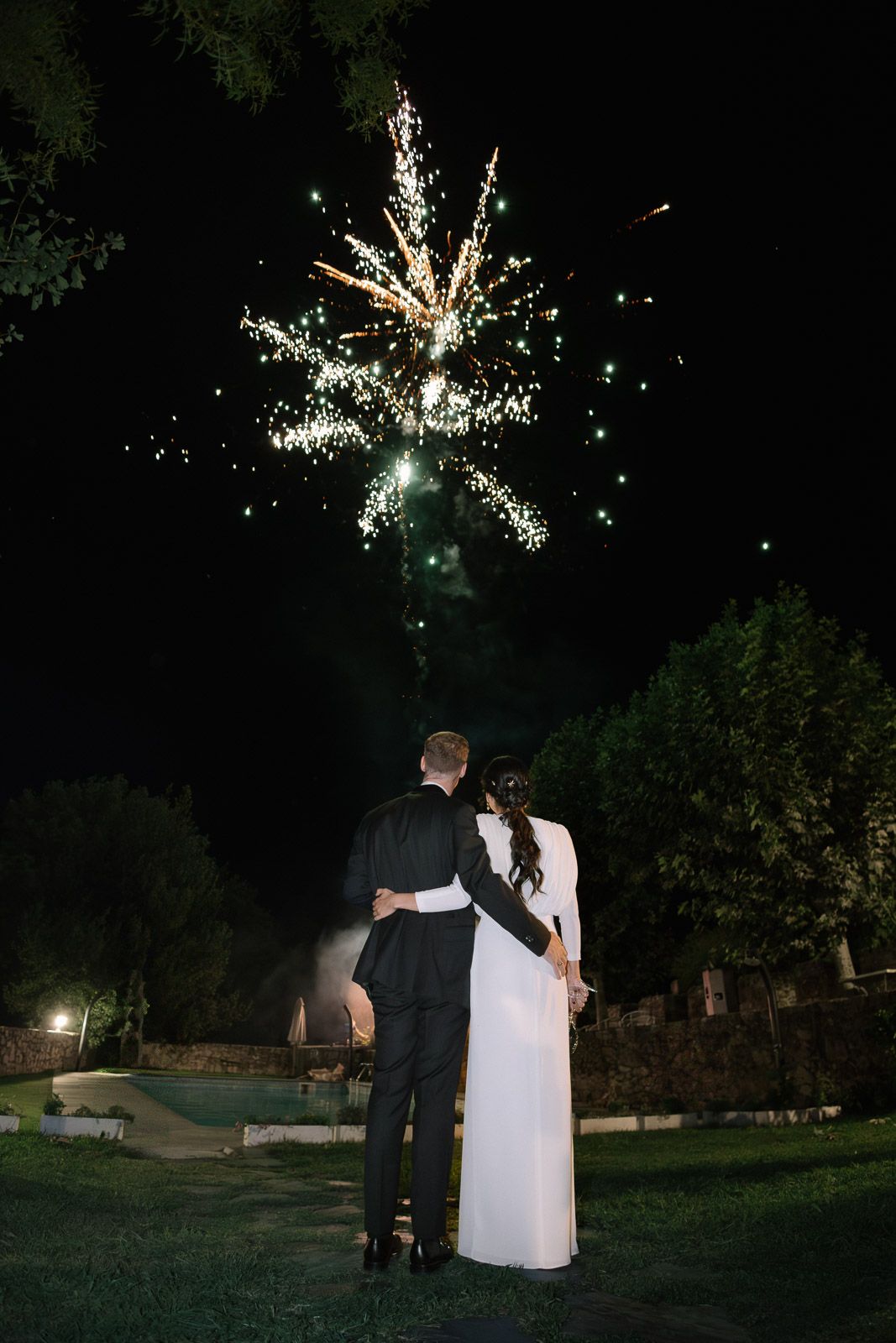 Bride and groom embrace while watching fireworks light up the night sky at their outdoor wedding celebration.