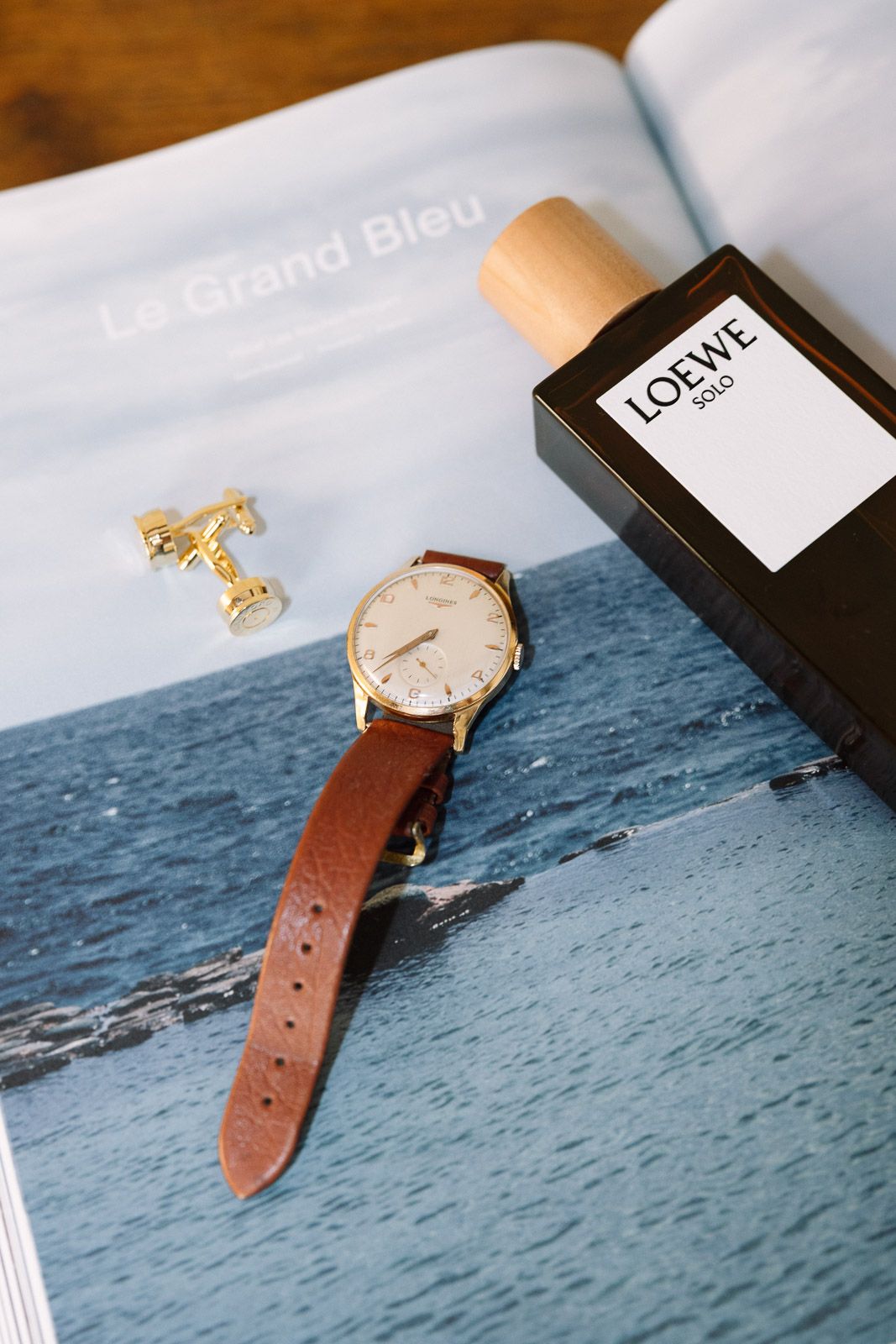 Gold watch with brown leather strap, Loewe Solo cologne, and gold cufflinks arranged on a magazine.