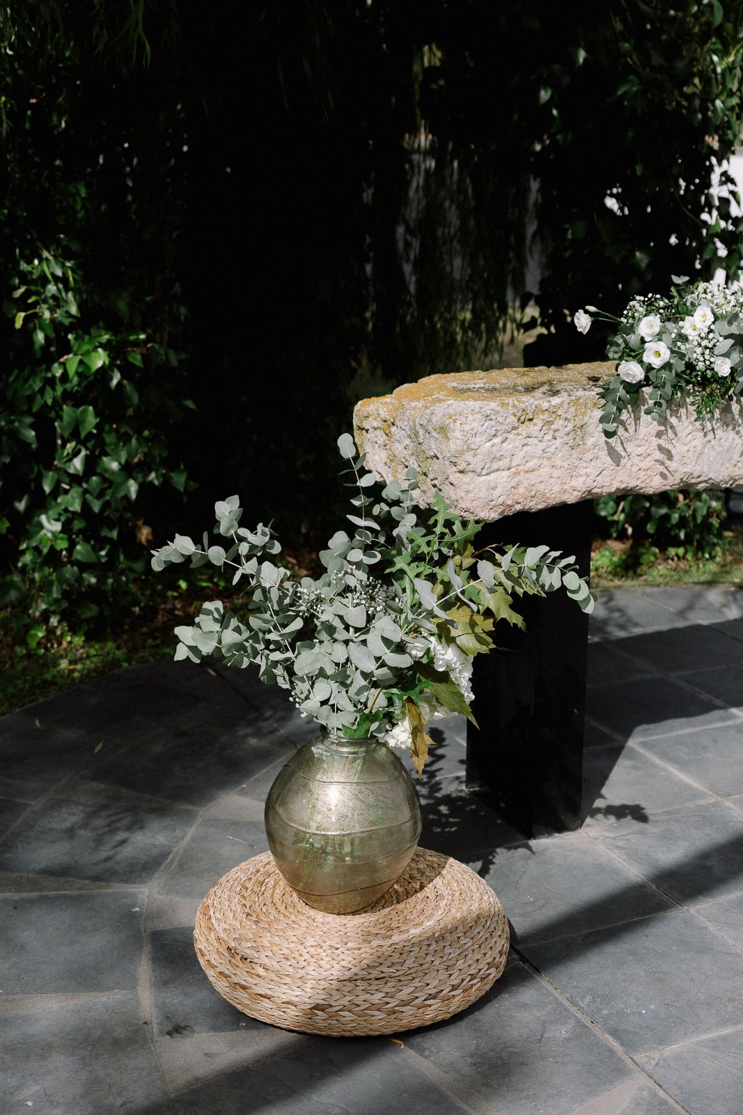 Rustic gold vase with eucalyptus and white flowers on woven mat beside stone bench outdoors.