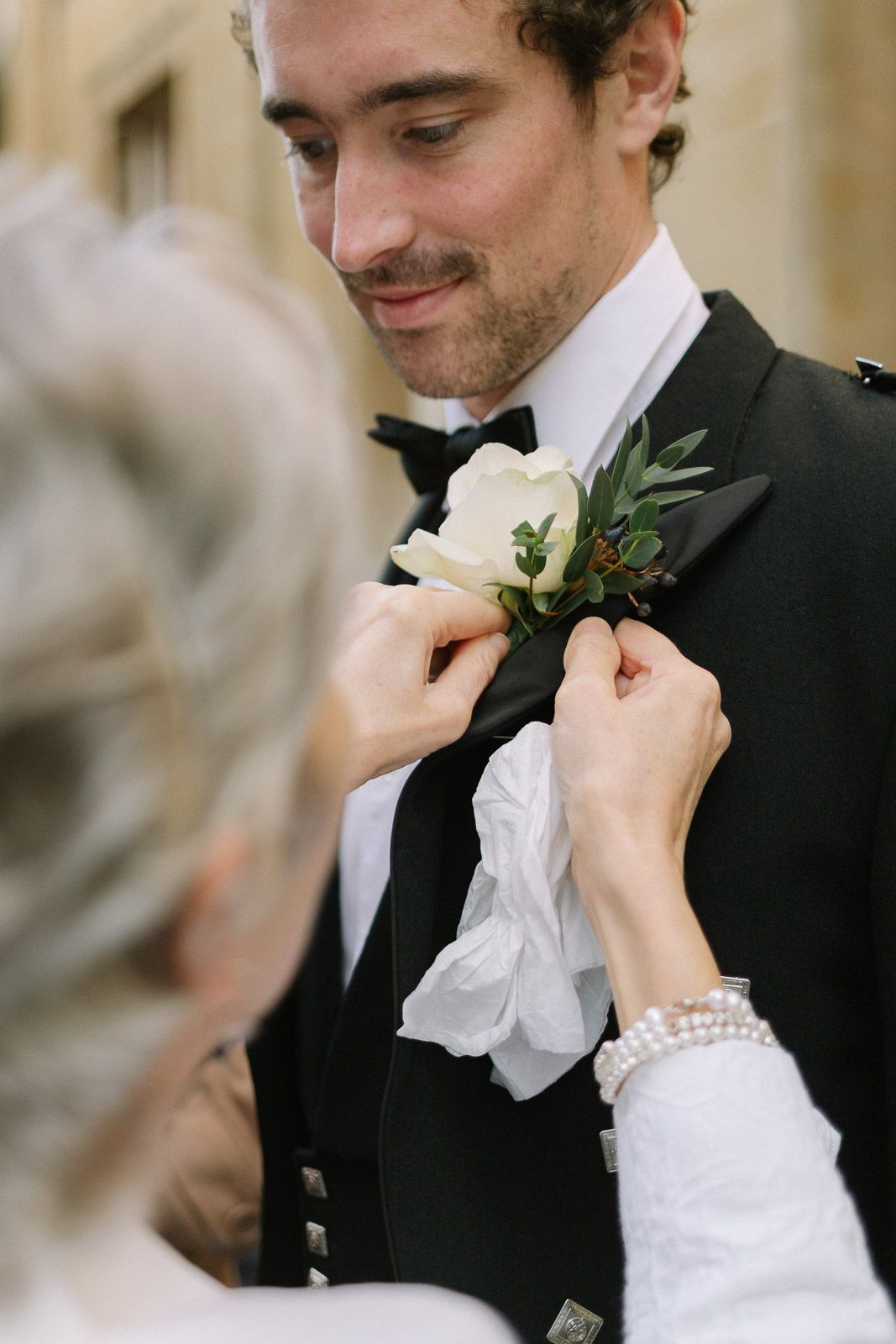 Woman pins white rose boutonniere on smiling groom in black tuxedo and bow tie before wedding ceremony.