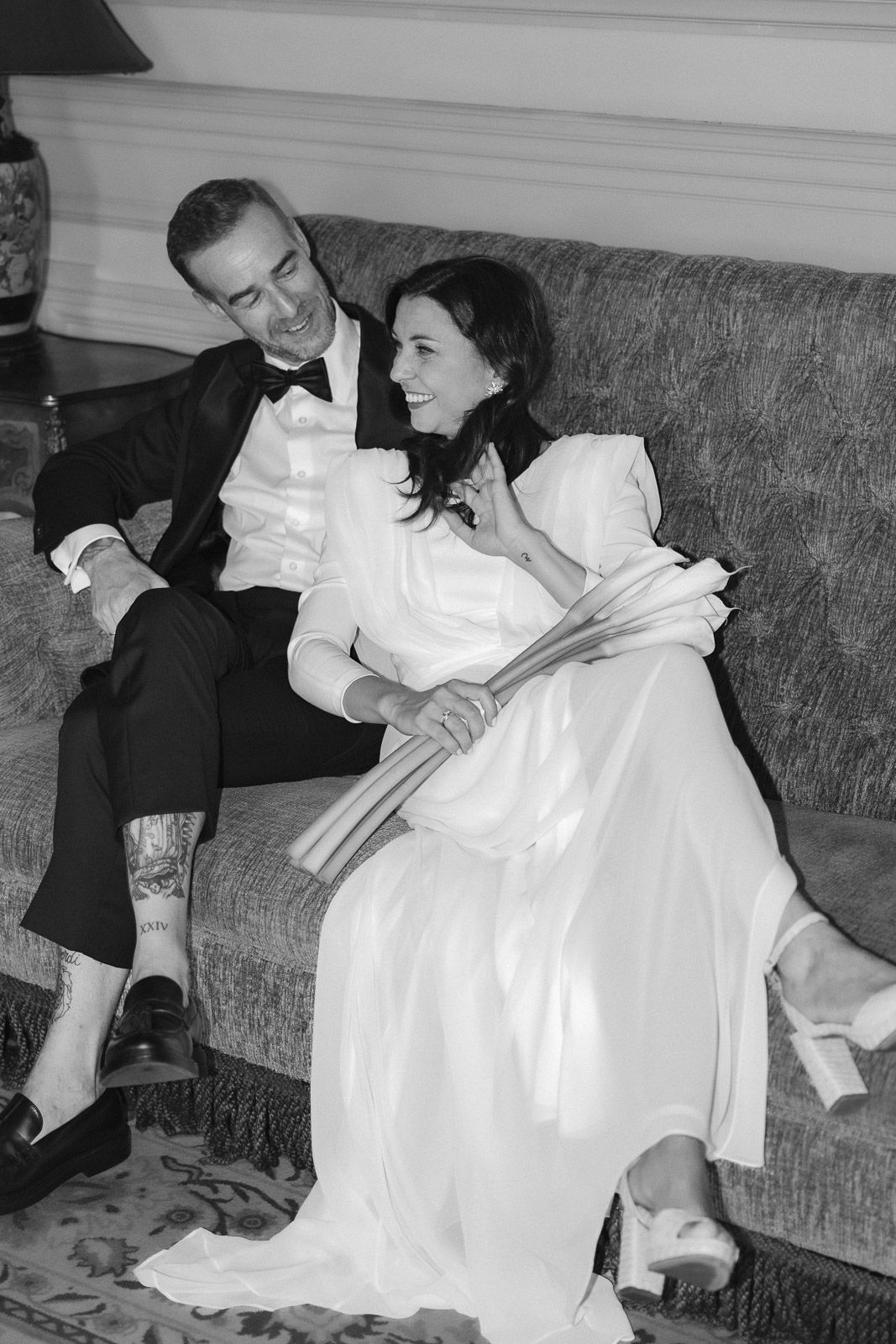 Black and white photo of bride and groom relaxing on sofa, bride holding bouquet in wedding dress.