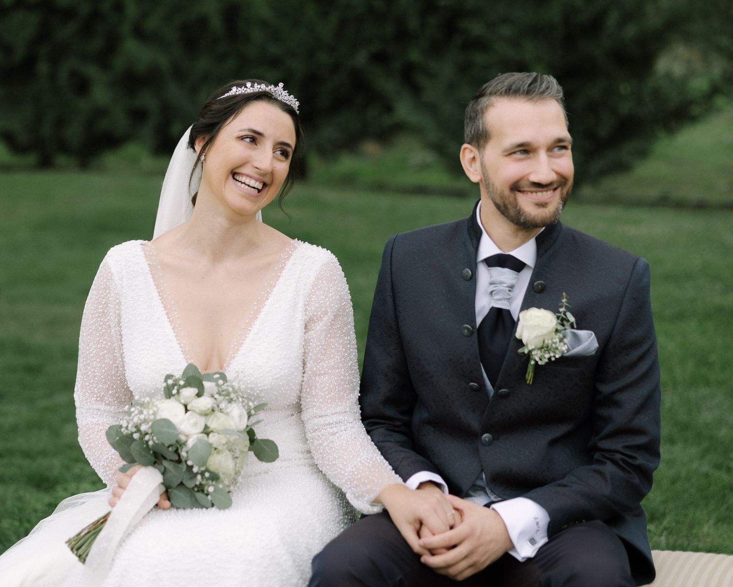 Happy bride and groom sitting together outdoors, bride holding white floral bouquet, groom wearing boutonniere.