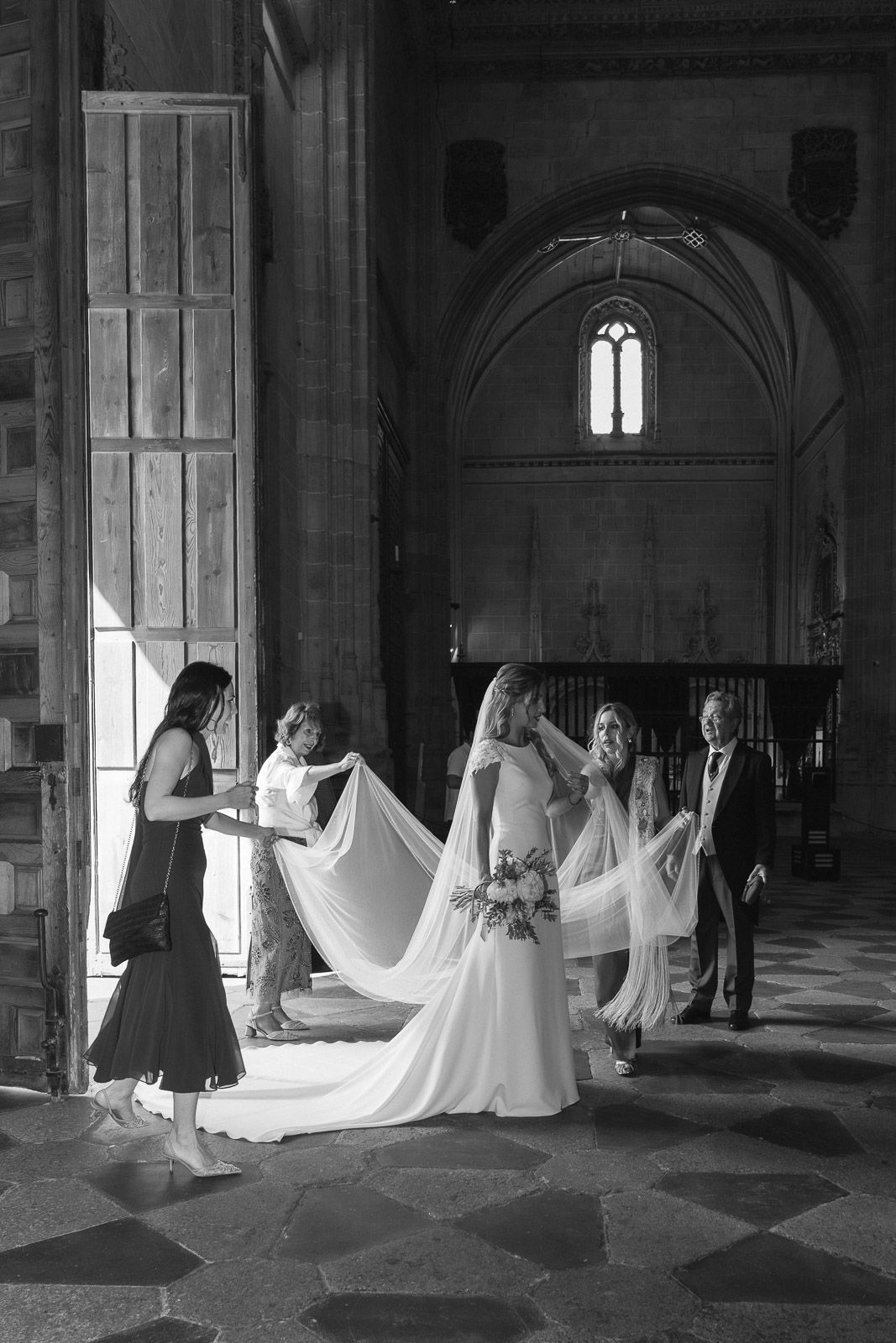 Black and white photo of a bride in a church with attendants arranging her veil before the wedding ceremony.