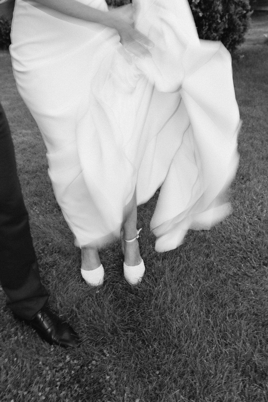 Black and white photo of bride in flowing white gown and heels standing beside groom in dress shoes on grass.