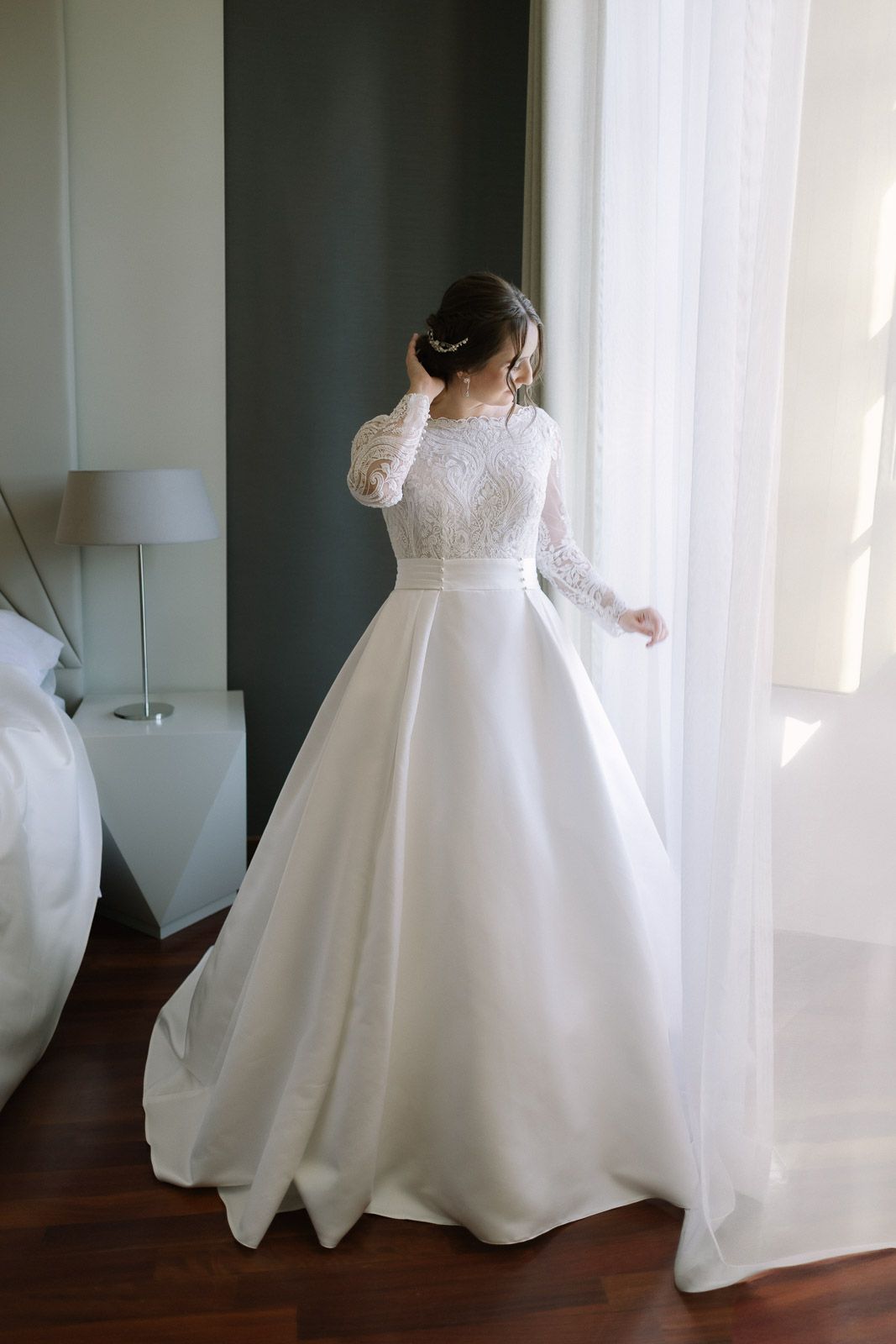 Bride in elegant long-sleeve lace ballgown wedding dress standing by a sunlit window in a hotel room.
