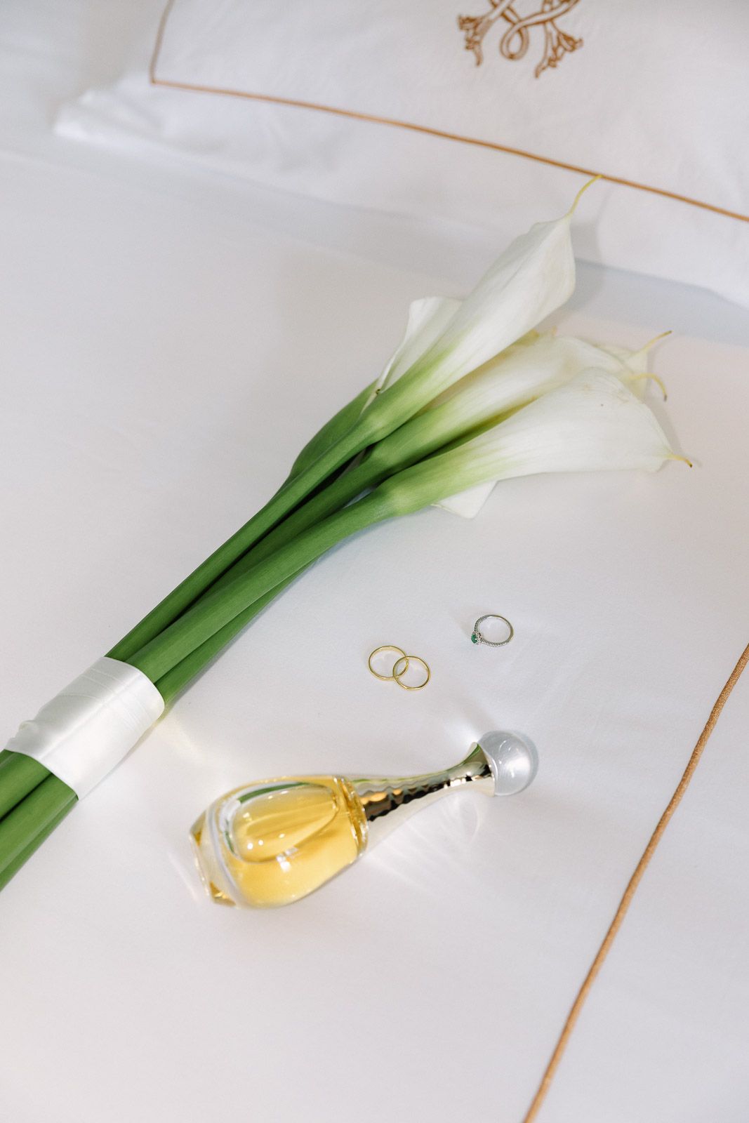 White calla lily bouquet with gold wedding rings and J'adore Dior perfume bottle on white linen.