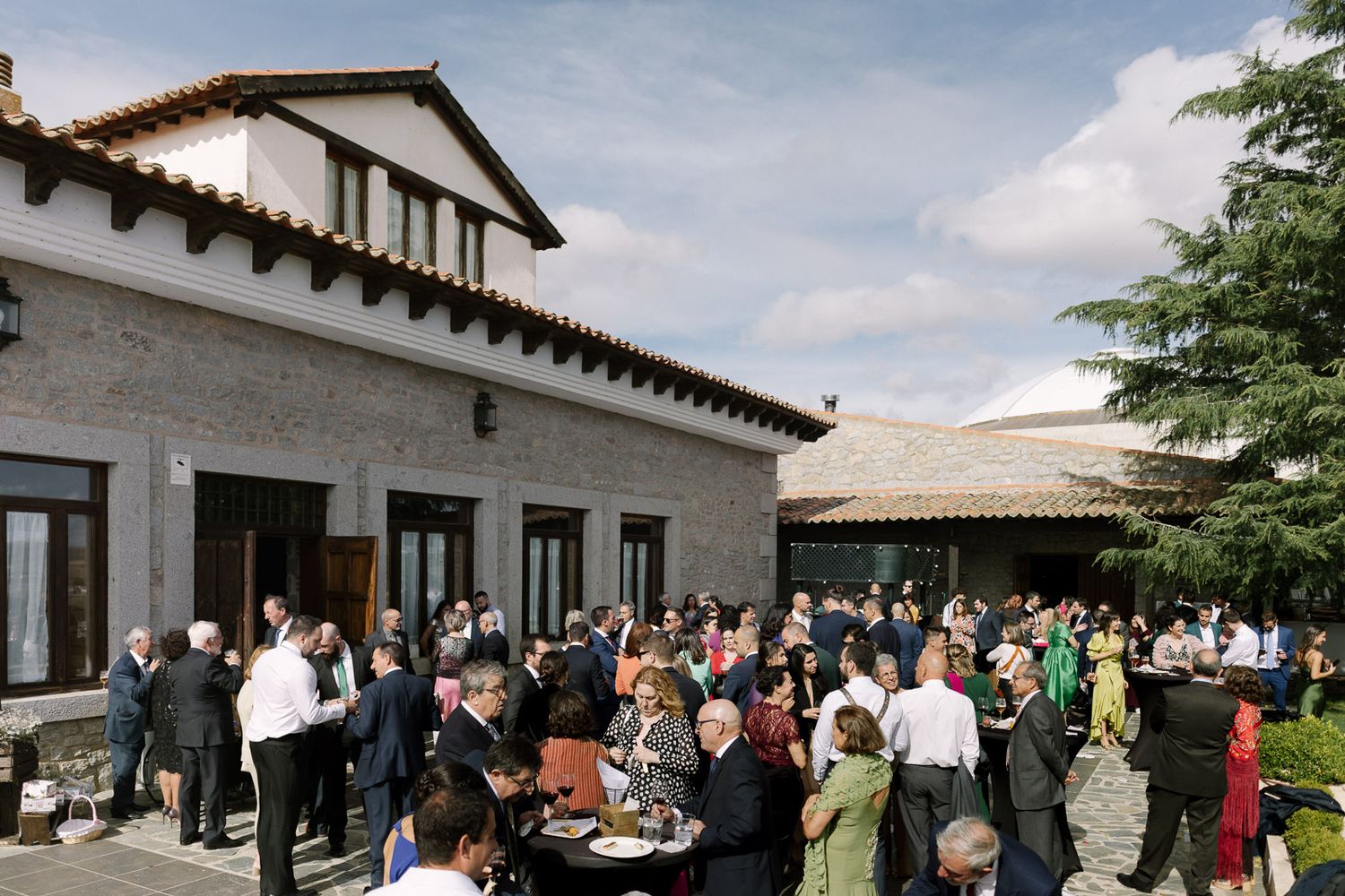 Elegant outdoor wedding reception at a rustic stone venue with guests mingling in a courtyard under a cloudy sky.