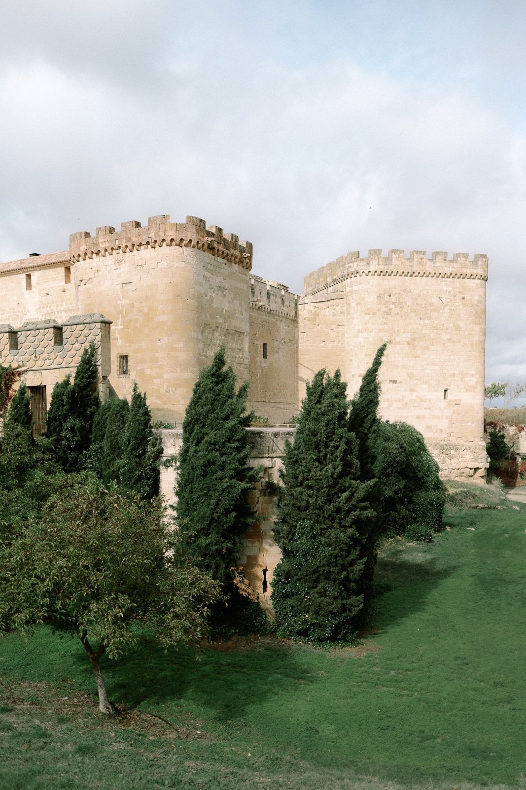 Medieval stone castle with crenellated towers surrounded by lush green cypress trees under a cloudy sky.