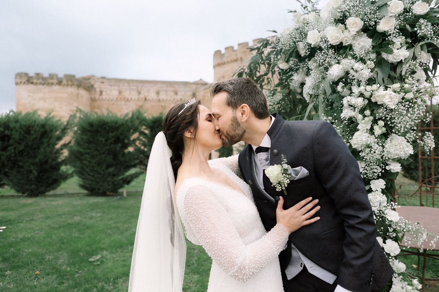 Bride and groom share first kiss at outdoor wedding ceremony with floral arch and historic castle backdrop.