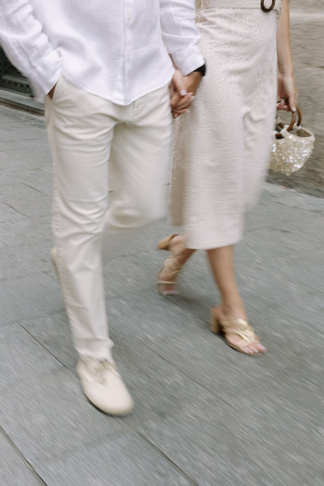 Couple holding hands while walking, wearing white and beige outfits with sandals on a gray pavement.