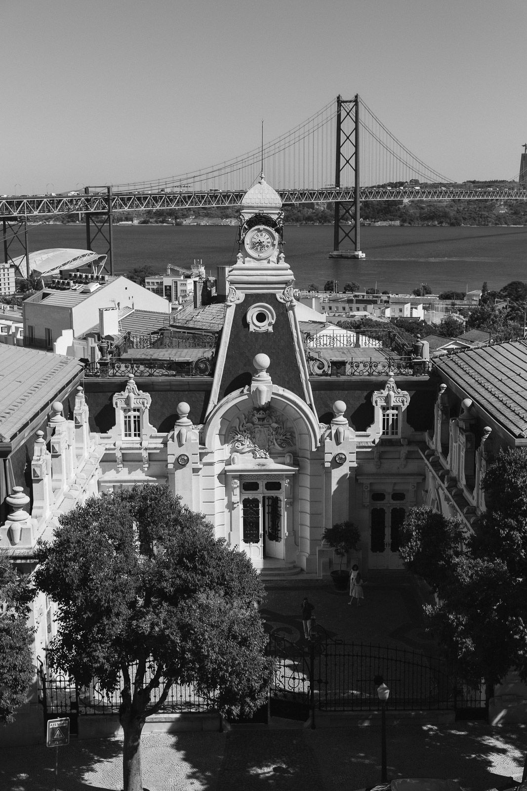 Black and white aerial view of ornate historic building with clock tower, Tagus River and 25 de Abril Bridge, Lisbon.