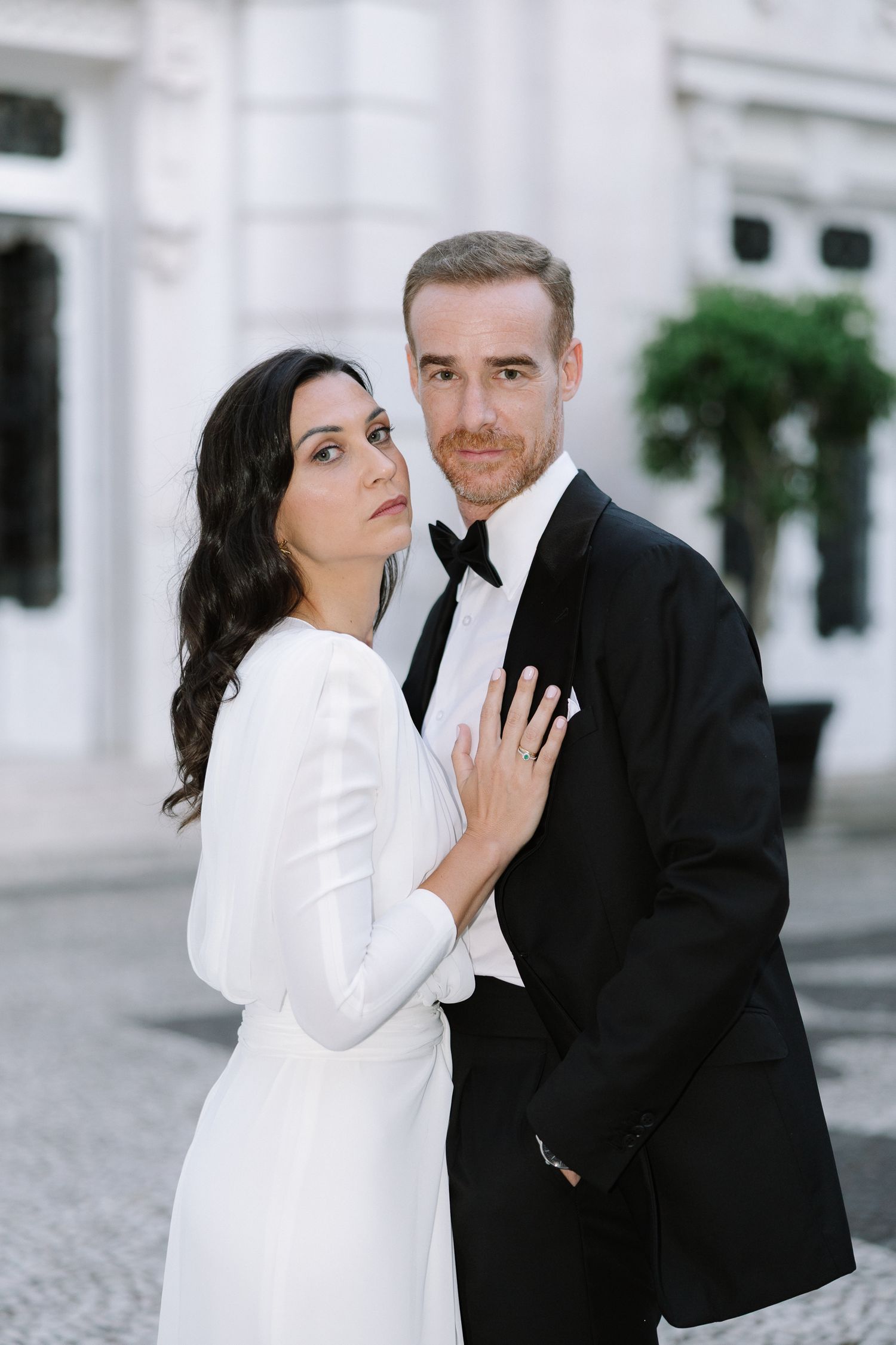 A couple in formal wedding attire shares intimate poses against a white architectural backdrop.